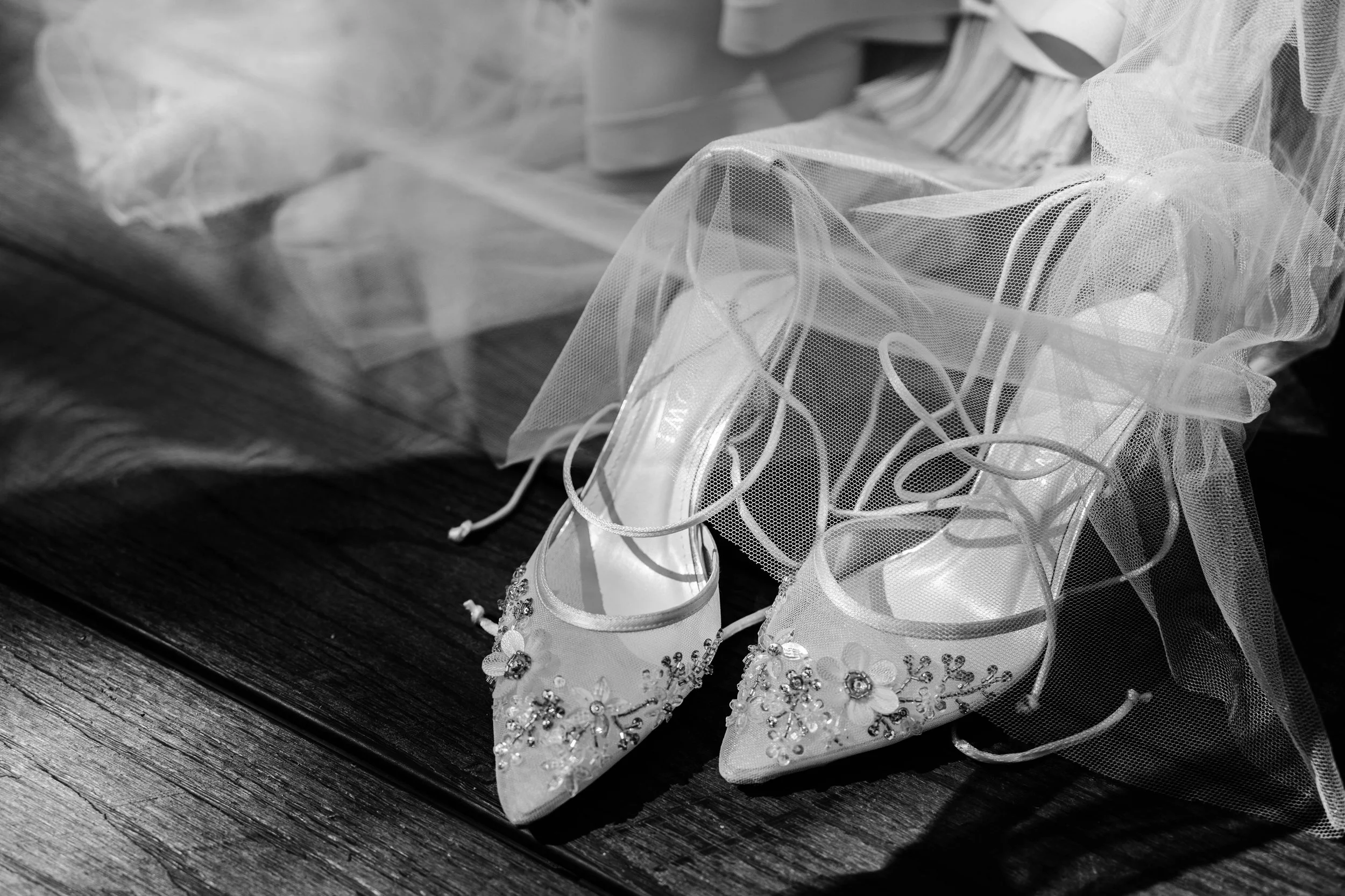 Elegant white bridal shoes with floral and rhinestone embellishments, placed on a dark wooden surface, partially covered by a tulle veil.