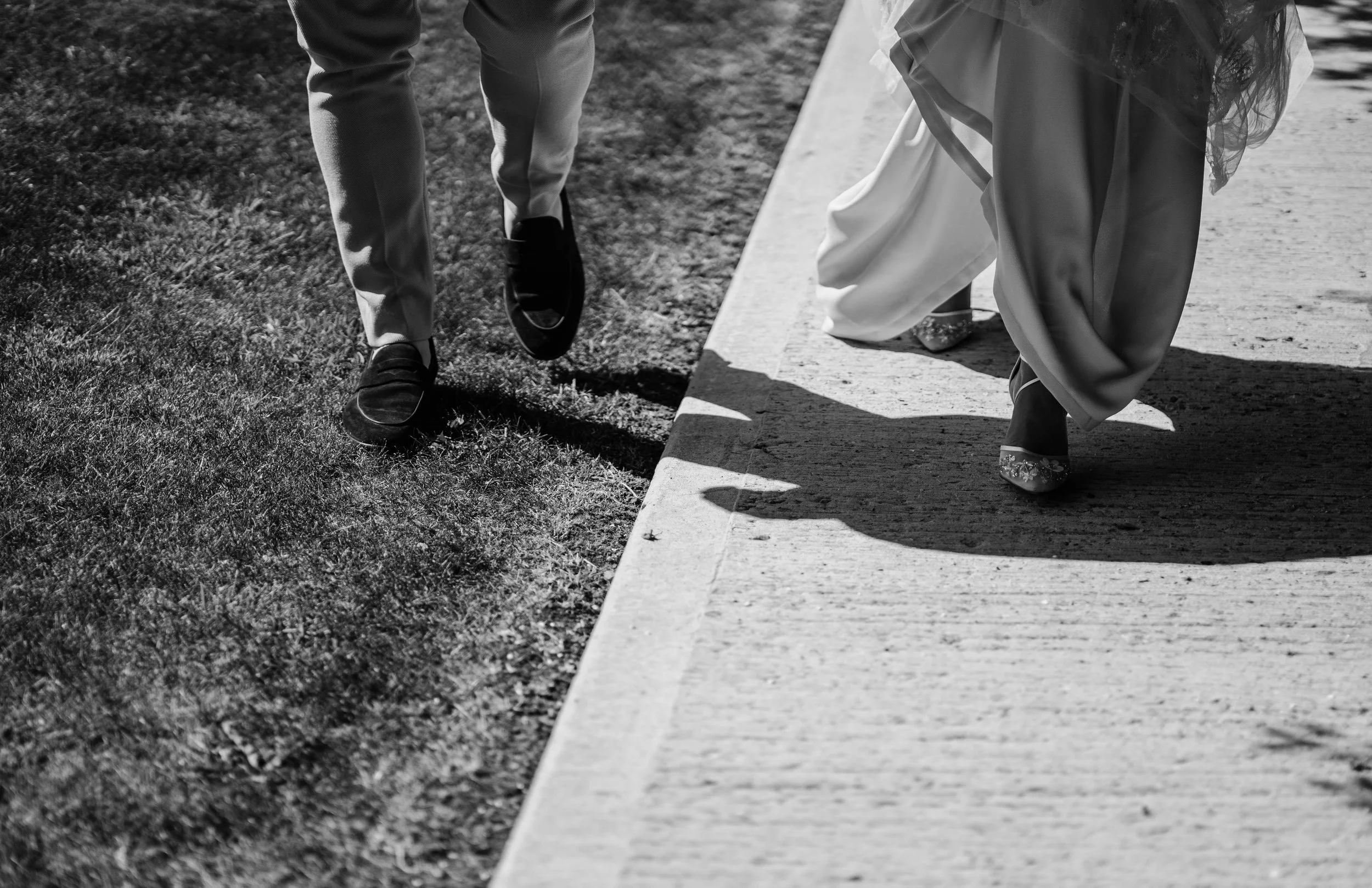 Close-up of a bride and groom walking together, focusing on their legs and shoes, with the bride wearing high heels and the groom in dress shoes, as they walk on a sidewalk.