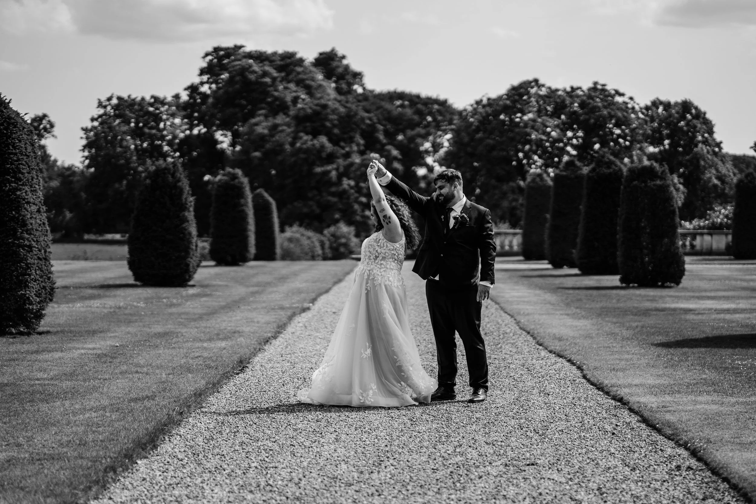 A black and white photo of a bride and groom dancing outdoors on a gravel path surrounded by tall, shaped bushes and large trees in the background.
