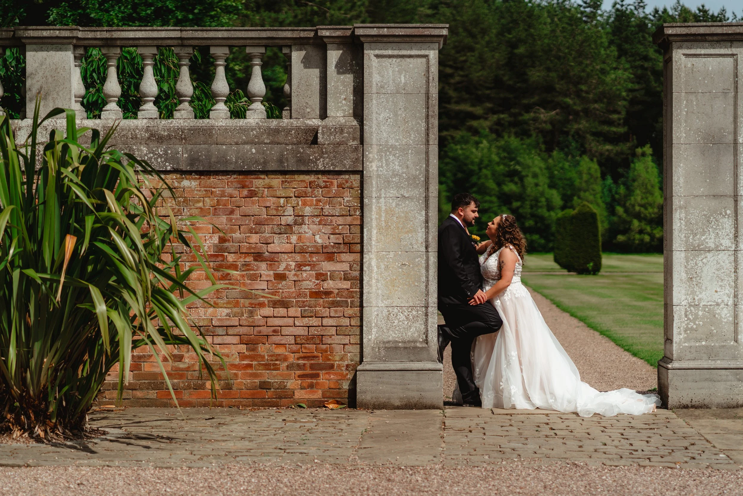 A bride and groom standing close together, holding hands, in a romantic pose outdoors. The groom is seated on a low stone wall, and the bride stands in front of him, gazing into his eyes. They are dressed in wedding attire, with the bride in a white 