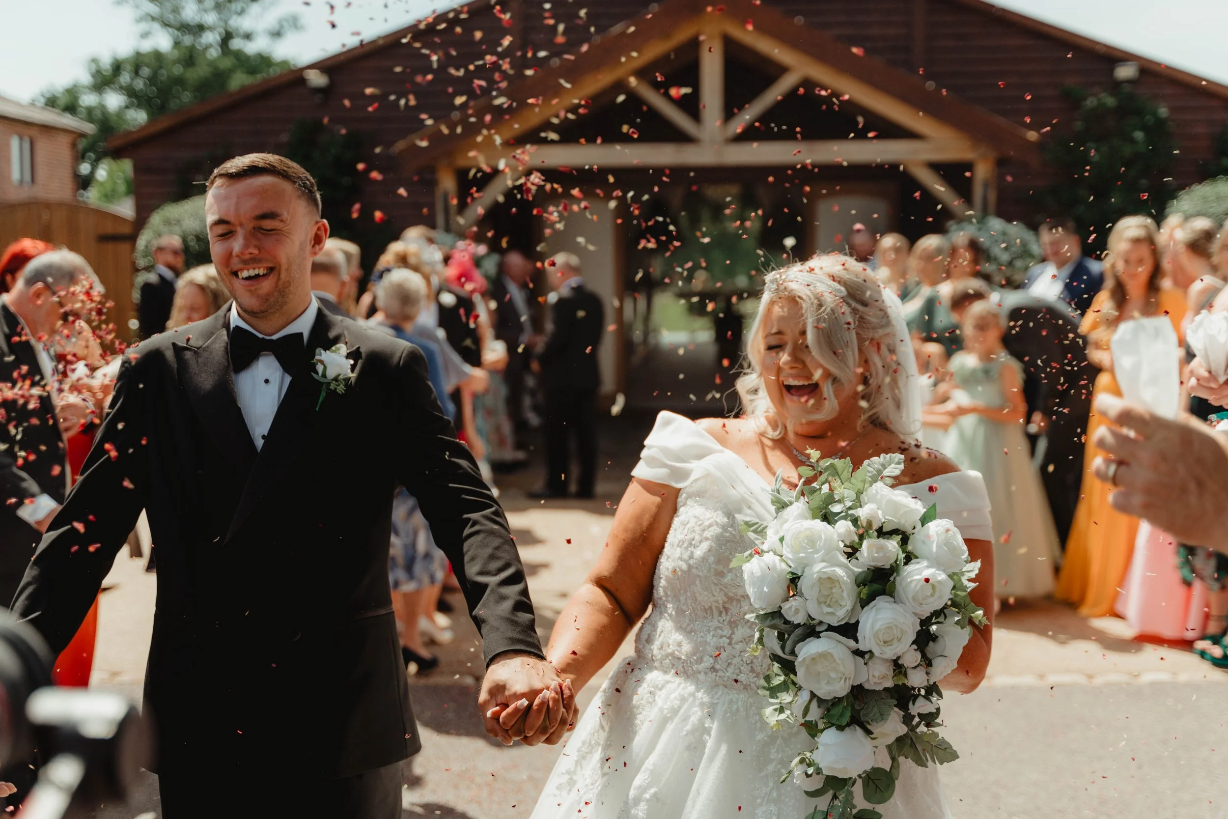 A newlywed couple holding hands, smiling and laughing as they walk away from their wedding ceremony, surrounded by friends and family throwing flower petals.