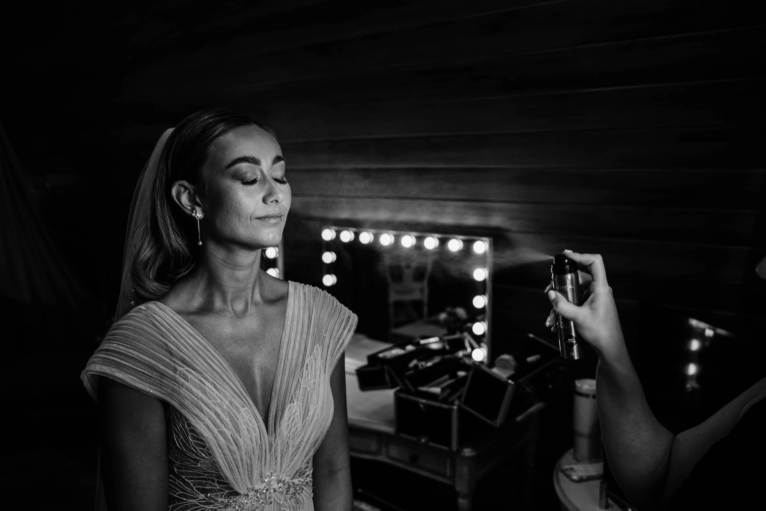 Bride with closed eyes in a wedding dress, while someone sprays her with a spray bottle, in a dressing room with lit mirror in the background.