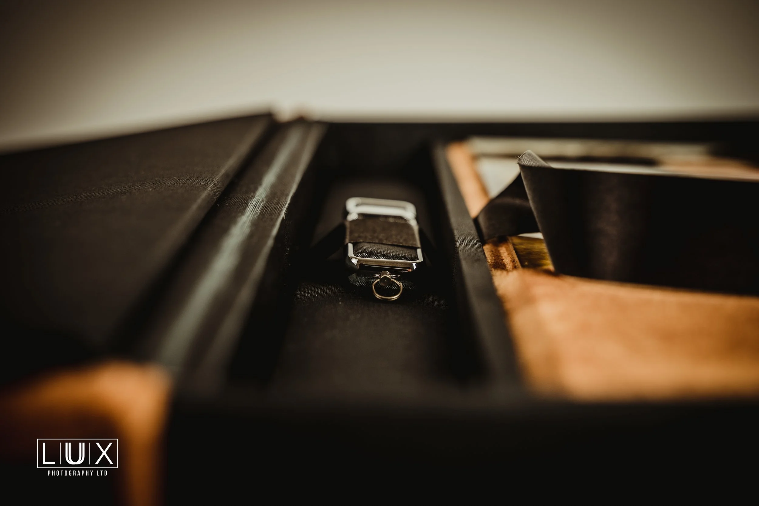 A black watch with a fabric strap and a metal ring attached, lying inside an open storage case with compartments, on a wooden surface.