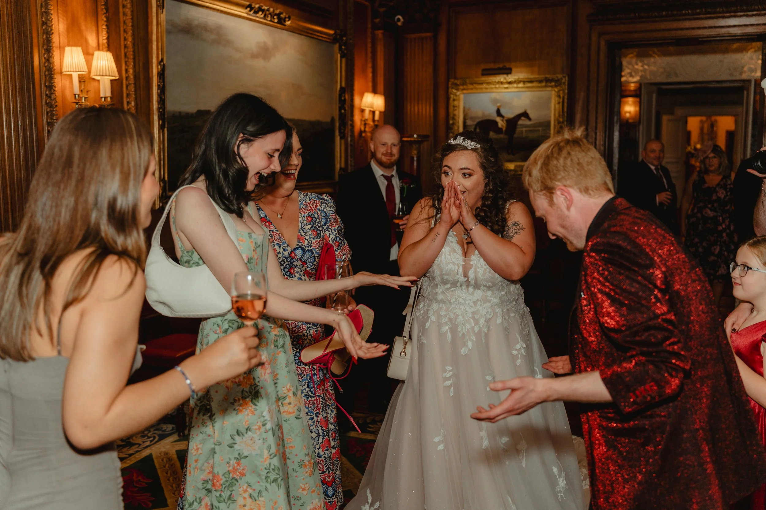 Group of people celebrating at a wedding reception, with a bride in a white gown and tiara covering her mouth in surprise, others smiling and clapping, in a room with wooden paneling and framed paintings.