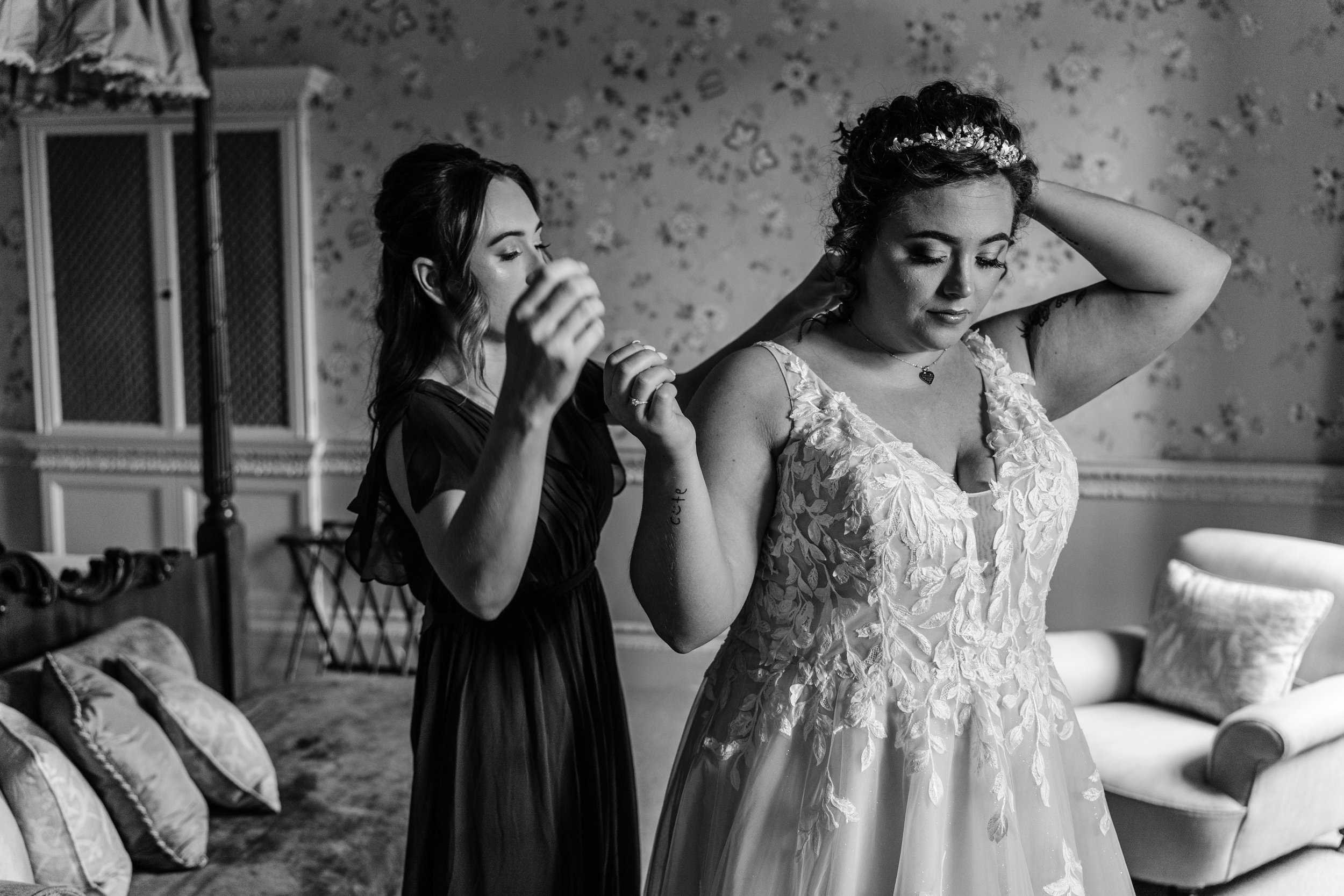 A bride in a lace wedding dress adjusting her hair, while a bridesmaid helps her in a living room with floral wallpaper and vintage furniture.