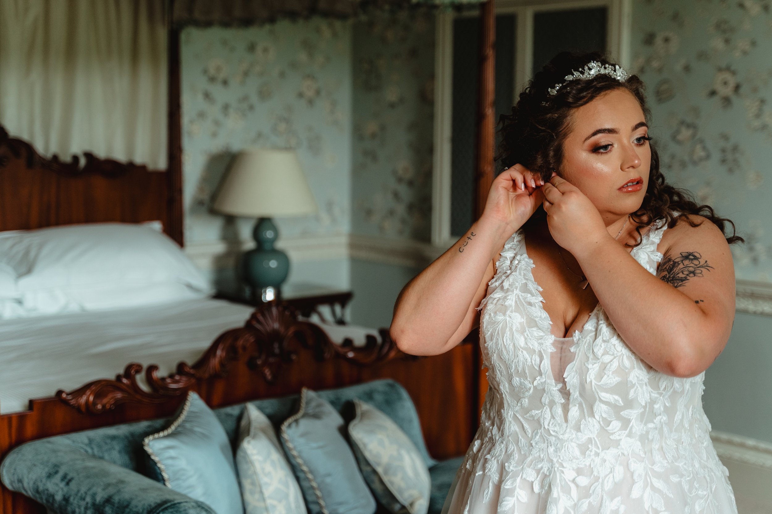 A woman in a wedding dress with dark curly hair and a tiara, standing in a bedroom, adjusting her earring.