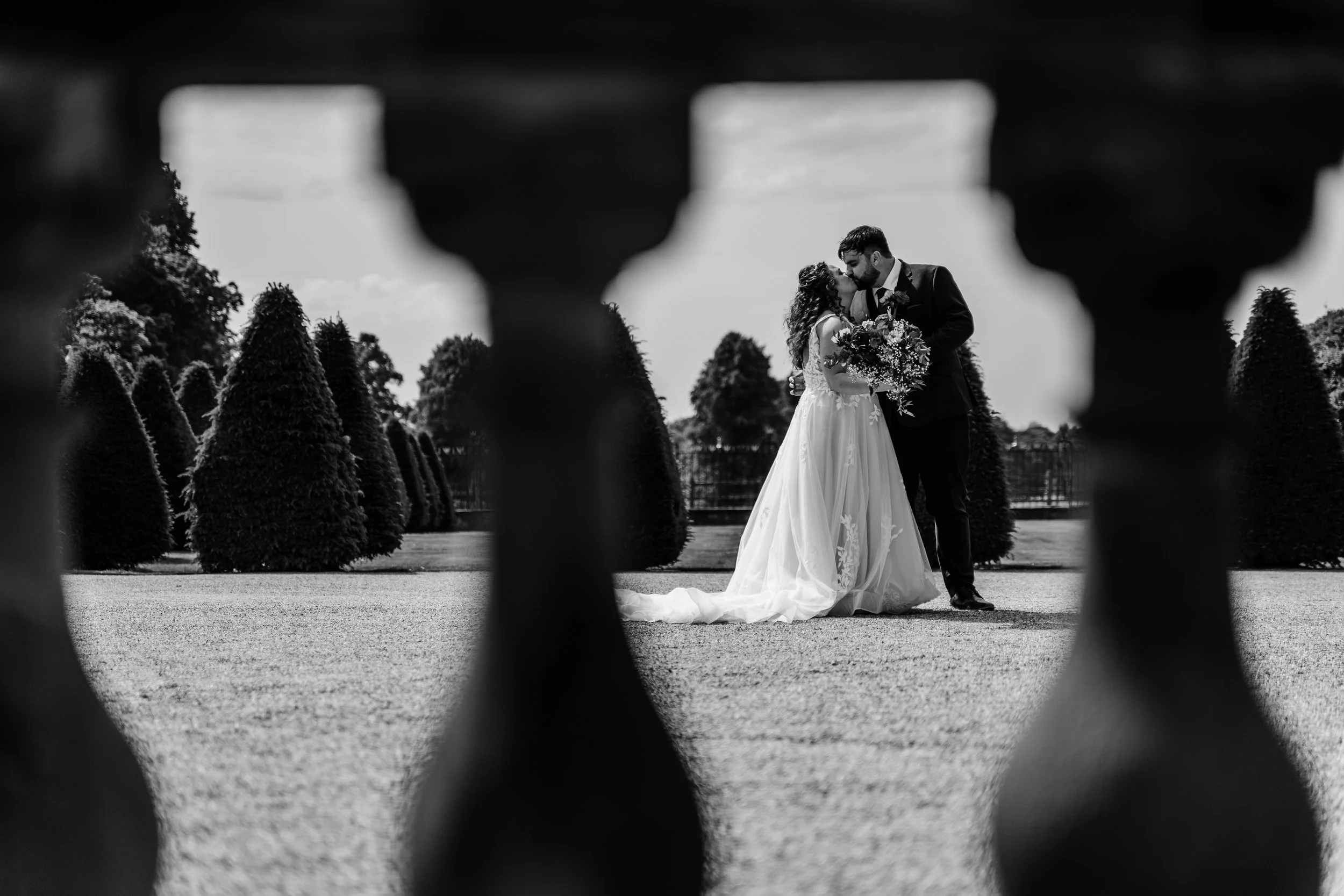 Black and white photo of a bride and groom kissing outdoors, seen through a decorative railing, with neatly trimmed bushes and trees in the background.