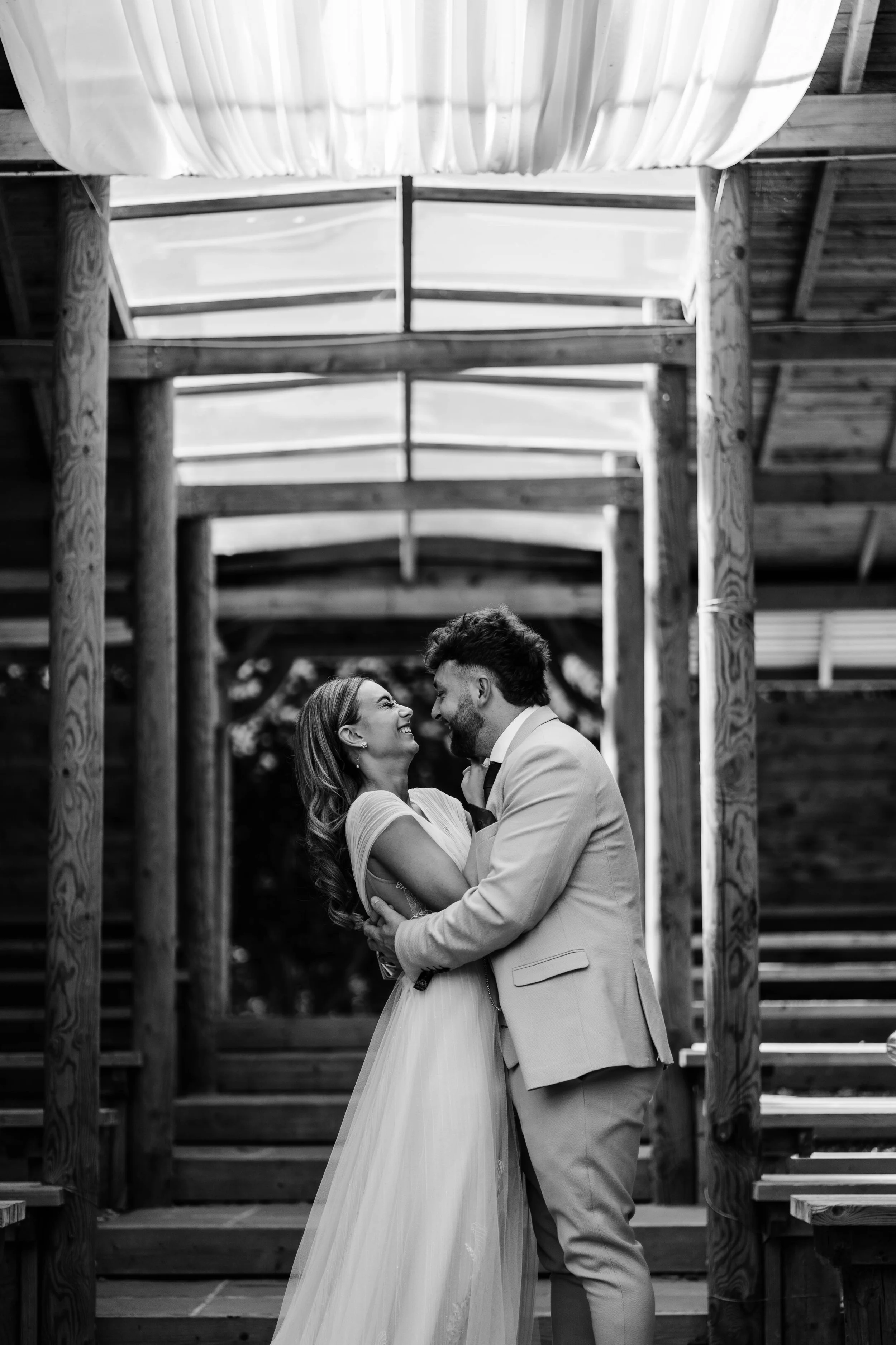 A couple in wedding attire smiling and embracing on a wooden staircase structure.