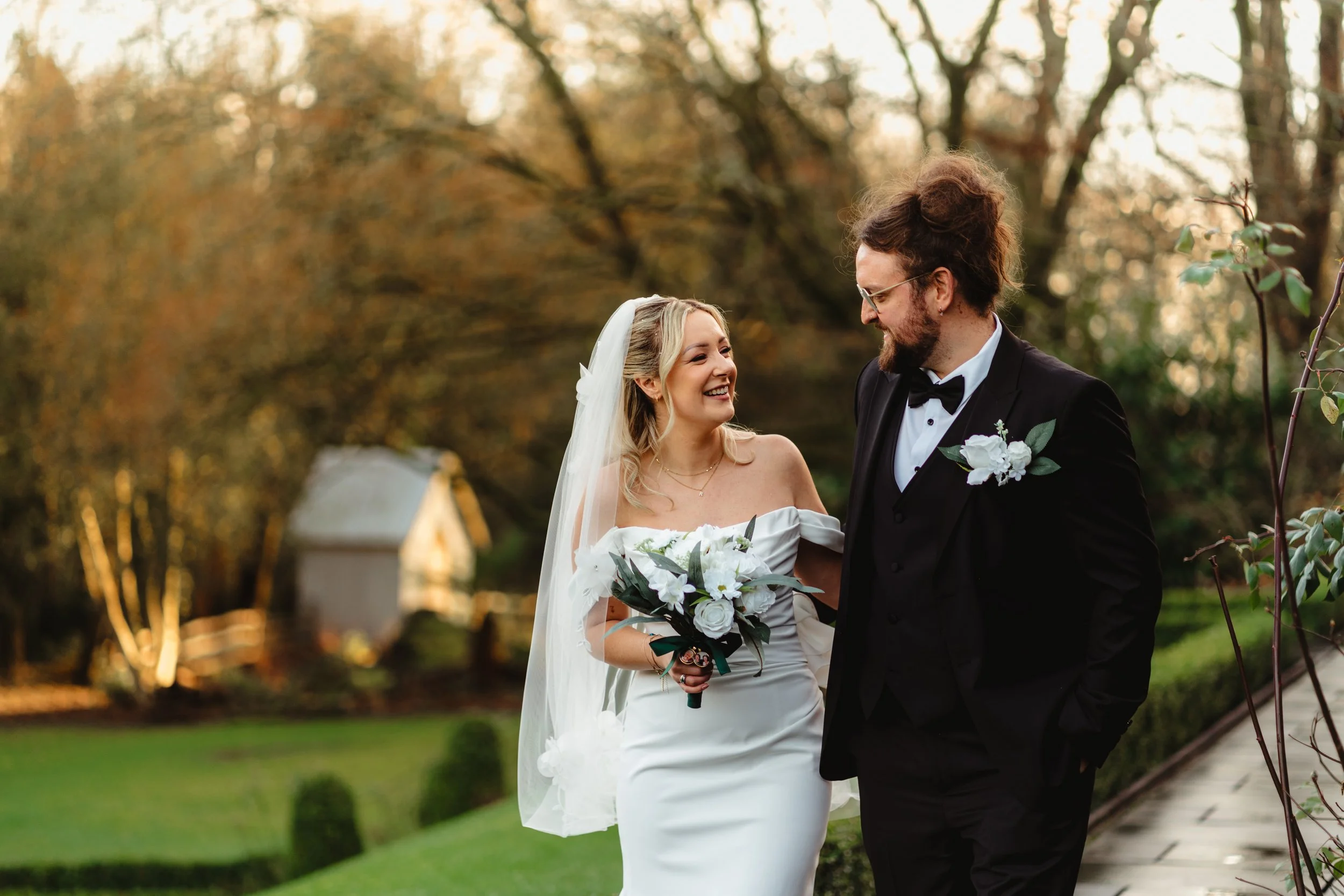 A bride and groom walking hand in hand through a stone archway, smiling and laughing during their wedding. The bride is holding a bouquet and wearing a white wedding dress, while the groom is dressed in a black suit.
