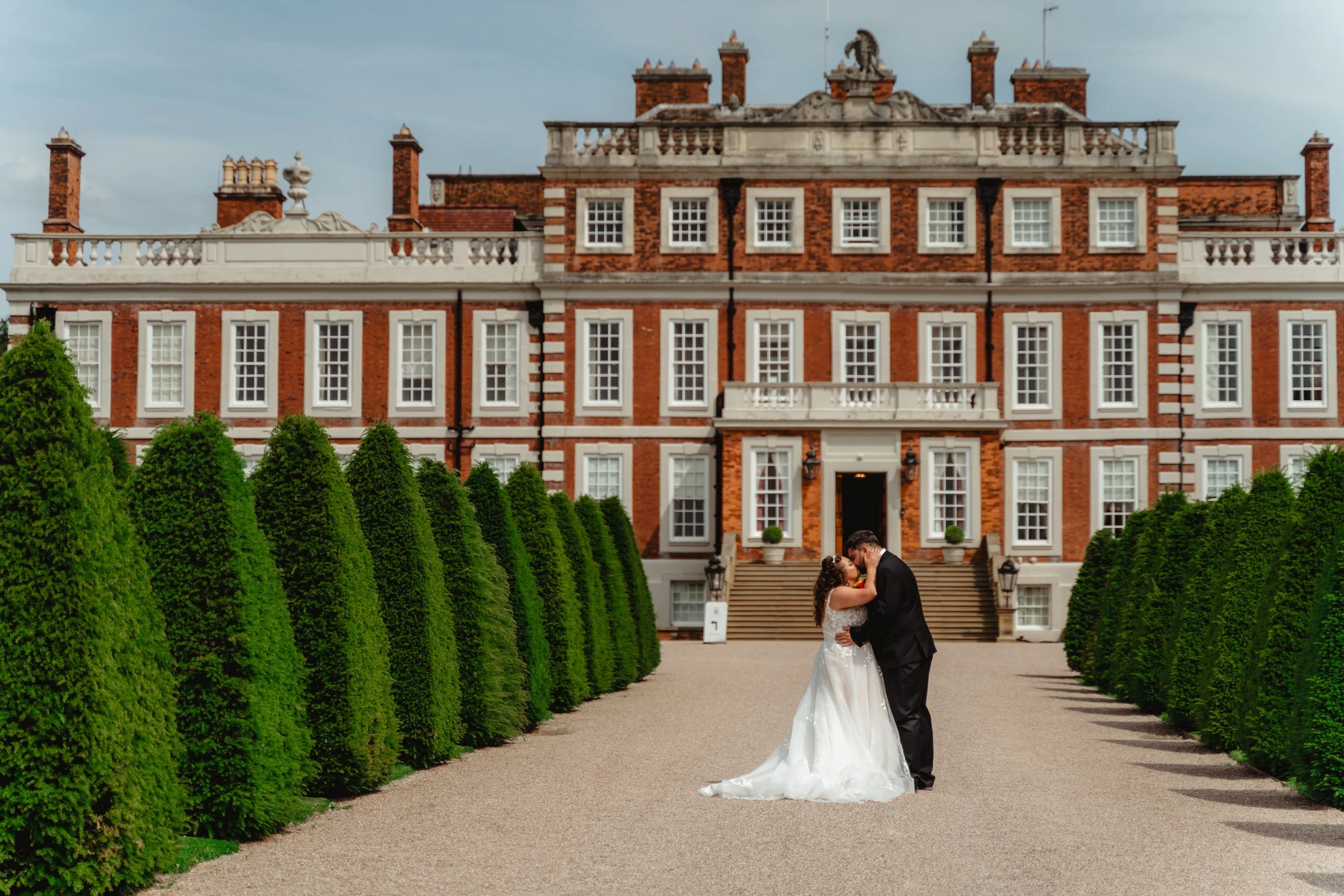 A bride and groom kissing in front of a large historic mansion, with symmetrical green hedges lining a gravel pathway.