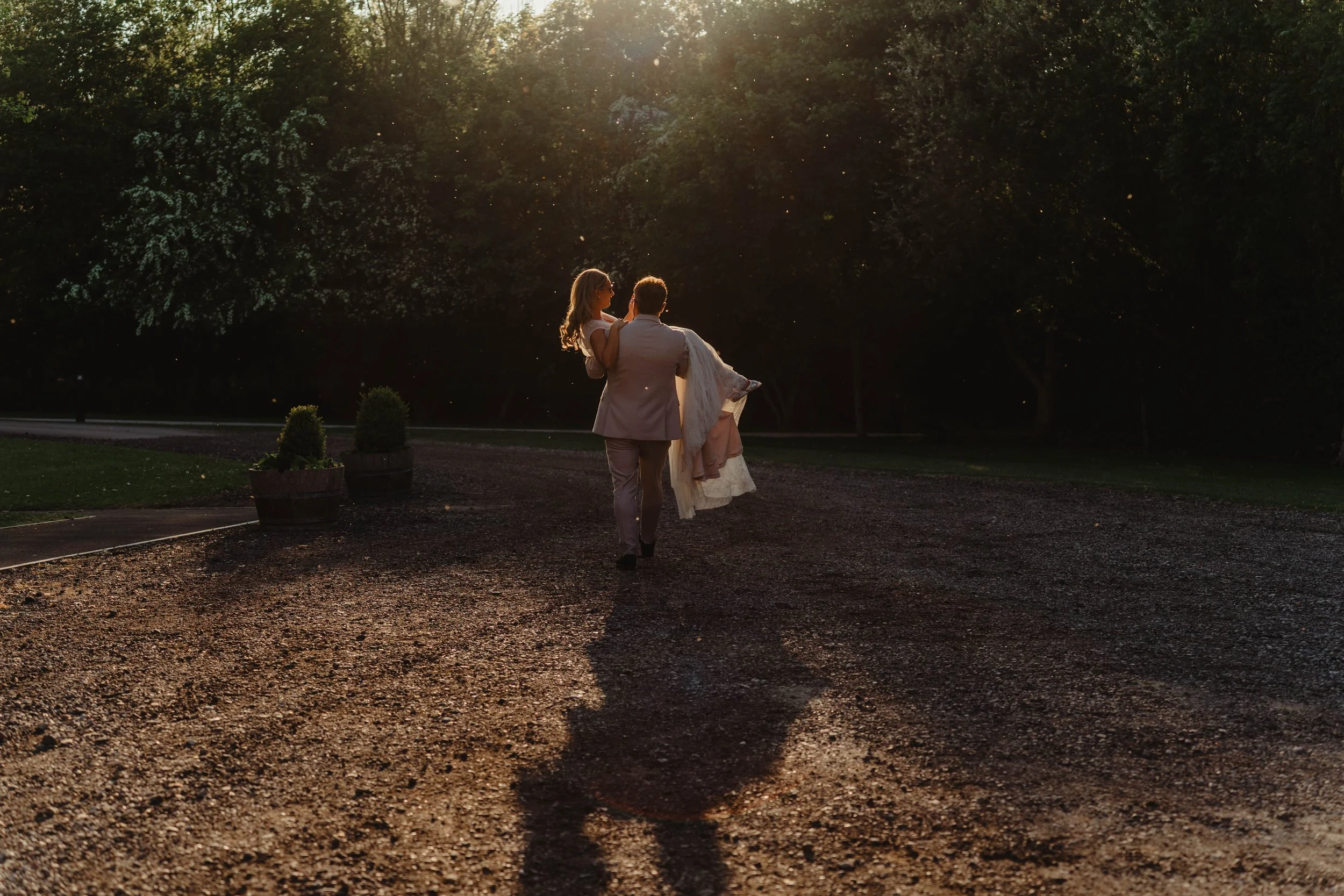A couple, with the man carrying the woman in his arms, walking on a gravel pathway in a park during sunset, with trees and potted plants in the background.