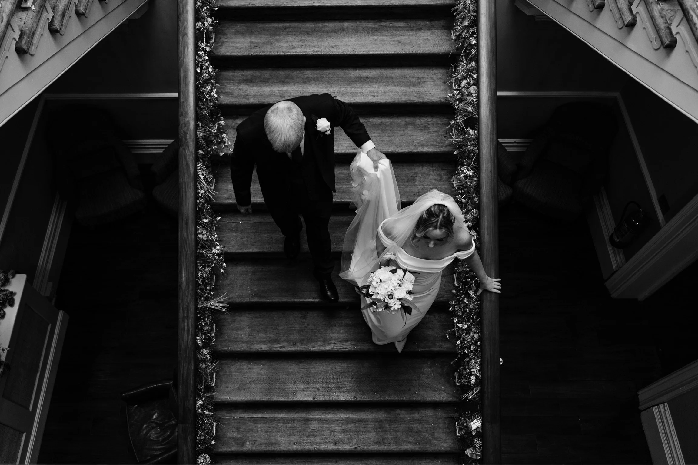 A black and white photo of a bride with a bouquet walking down a staircase, accompanied by a man in a suit, possibly her father, with the staircase decorated with tinsel for a wedding or special occasion.