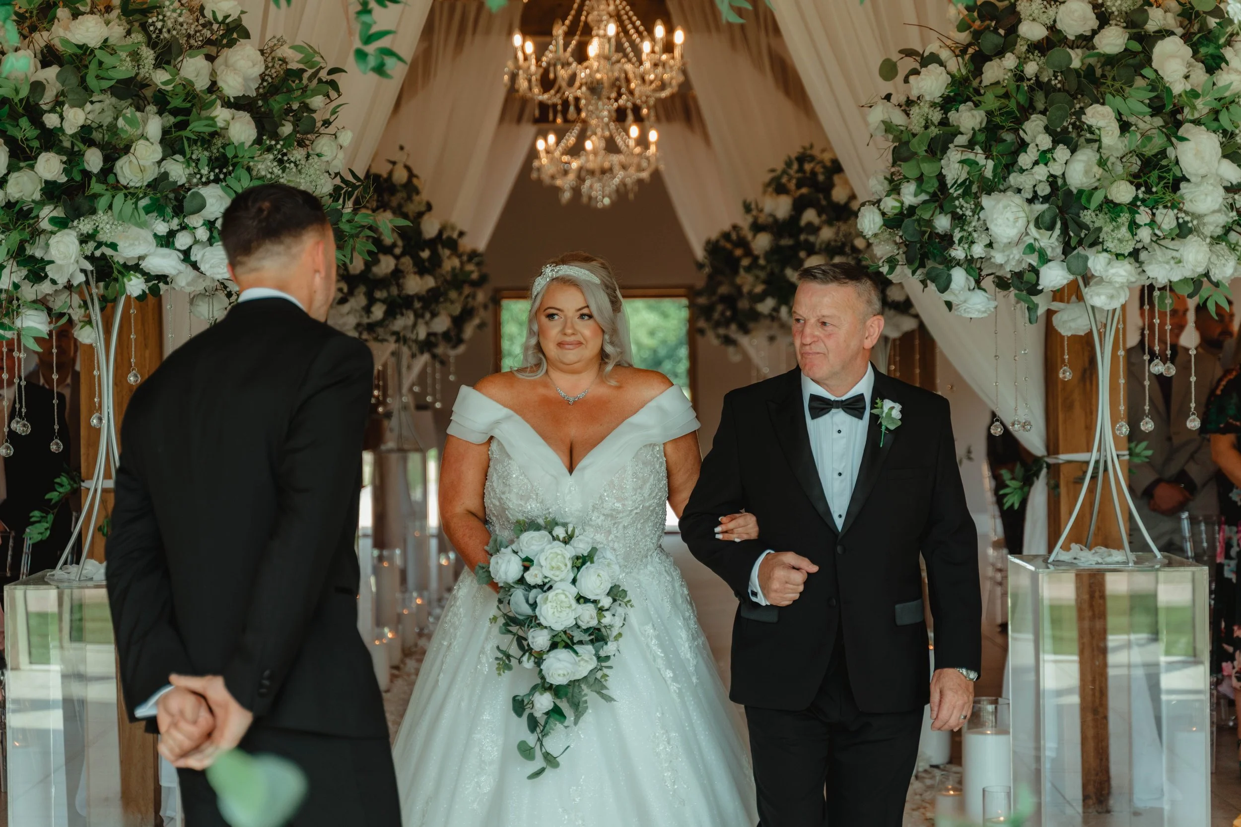A bride in a white wedding gown holding a bouquet of white roses and greenery, walking down the aisle with her father who is wearing a black tuxedo, during a wedding ceremony inside a decorated venue with floral arrangements and chandeliers.