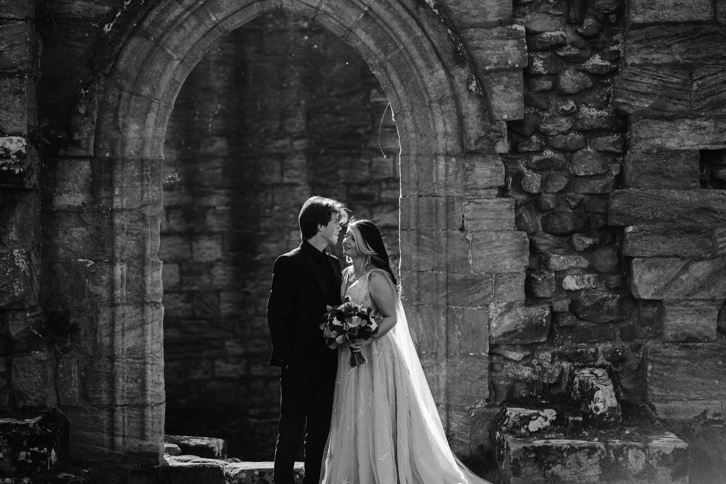 Black and white photo of a bride and groom standing close together under an ancient stone arch, with the bride holding a bouquet of flowers and looking at the groom.