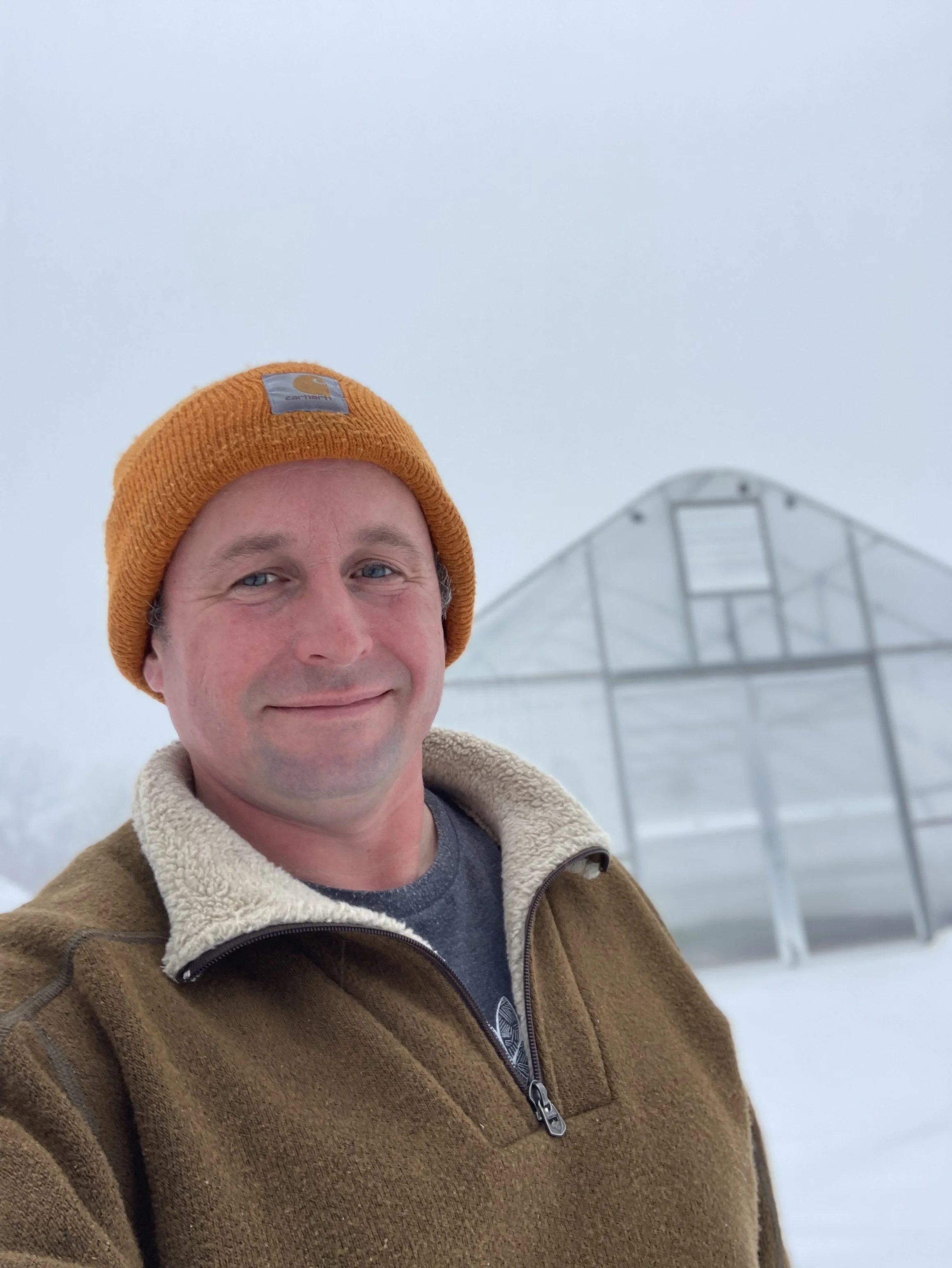 A smiling man in a brown jacket and orange beanie taking a selfie outdoors in a snowy landscape with a greenhouse in the background.