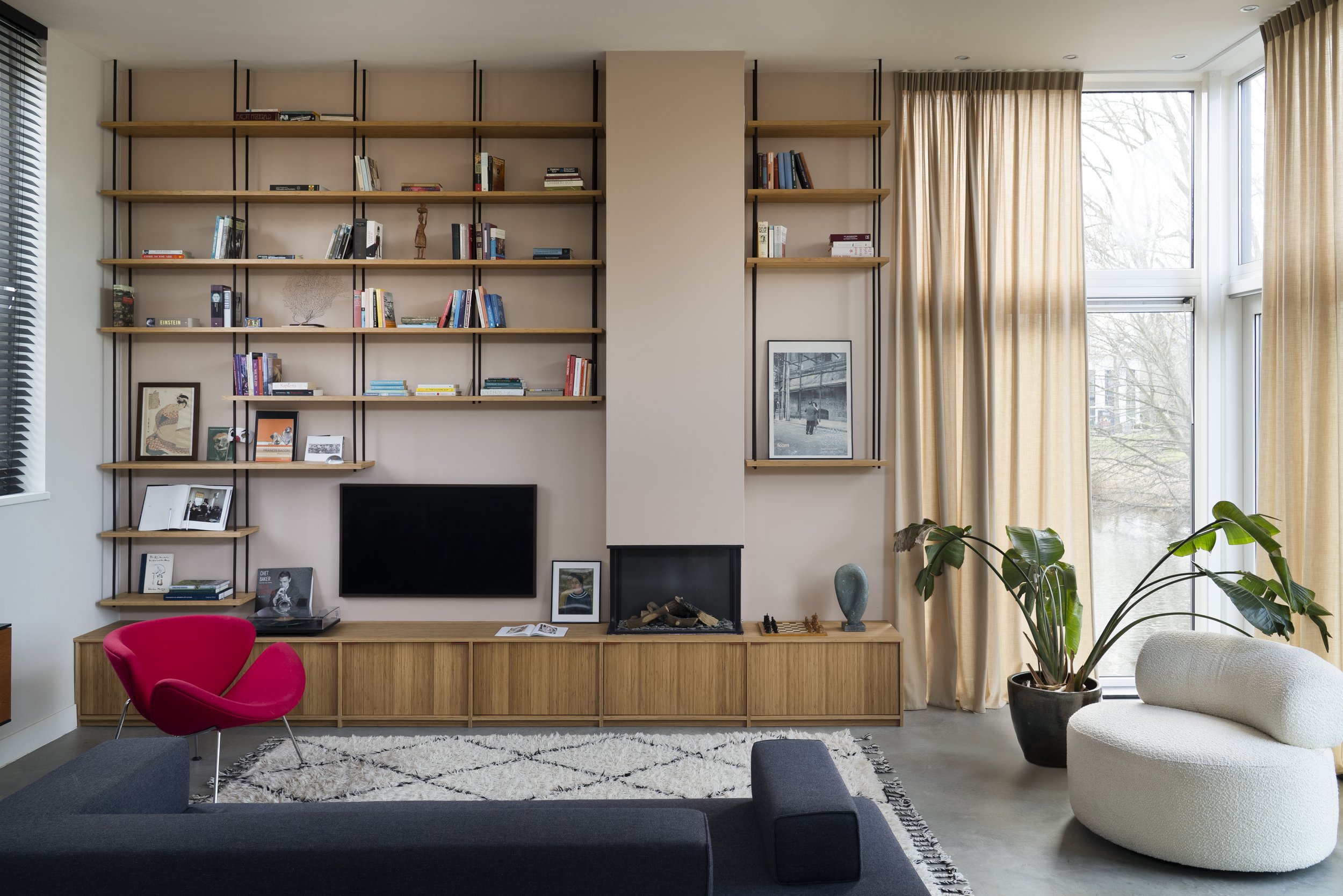 Modern living room with a large wall-mounted bookshelf filled with books and decorative items. A flat-screen TV is mounted on the wall, below the shelves. There is a bright pink chair, a white curved chair, and a black sofa. The room has large window