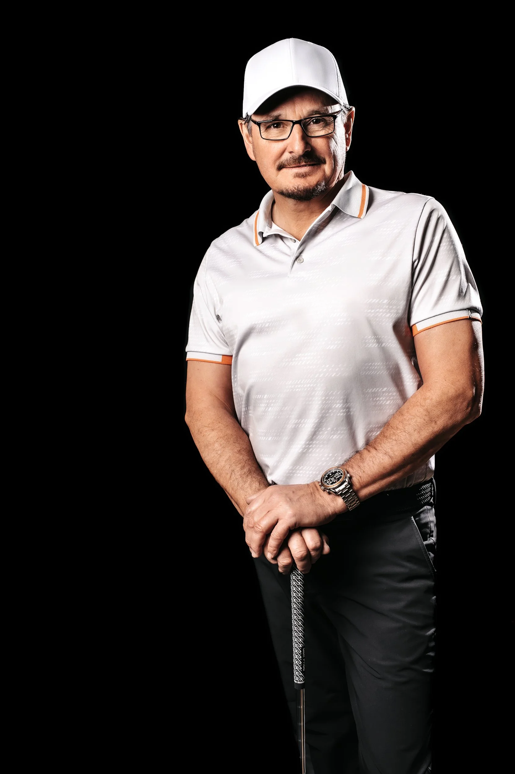 Man in white polo shirt and cap holding a golf club against black background