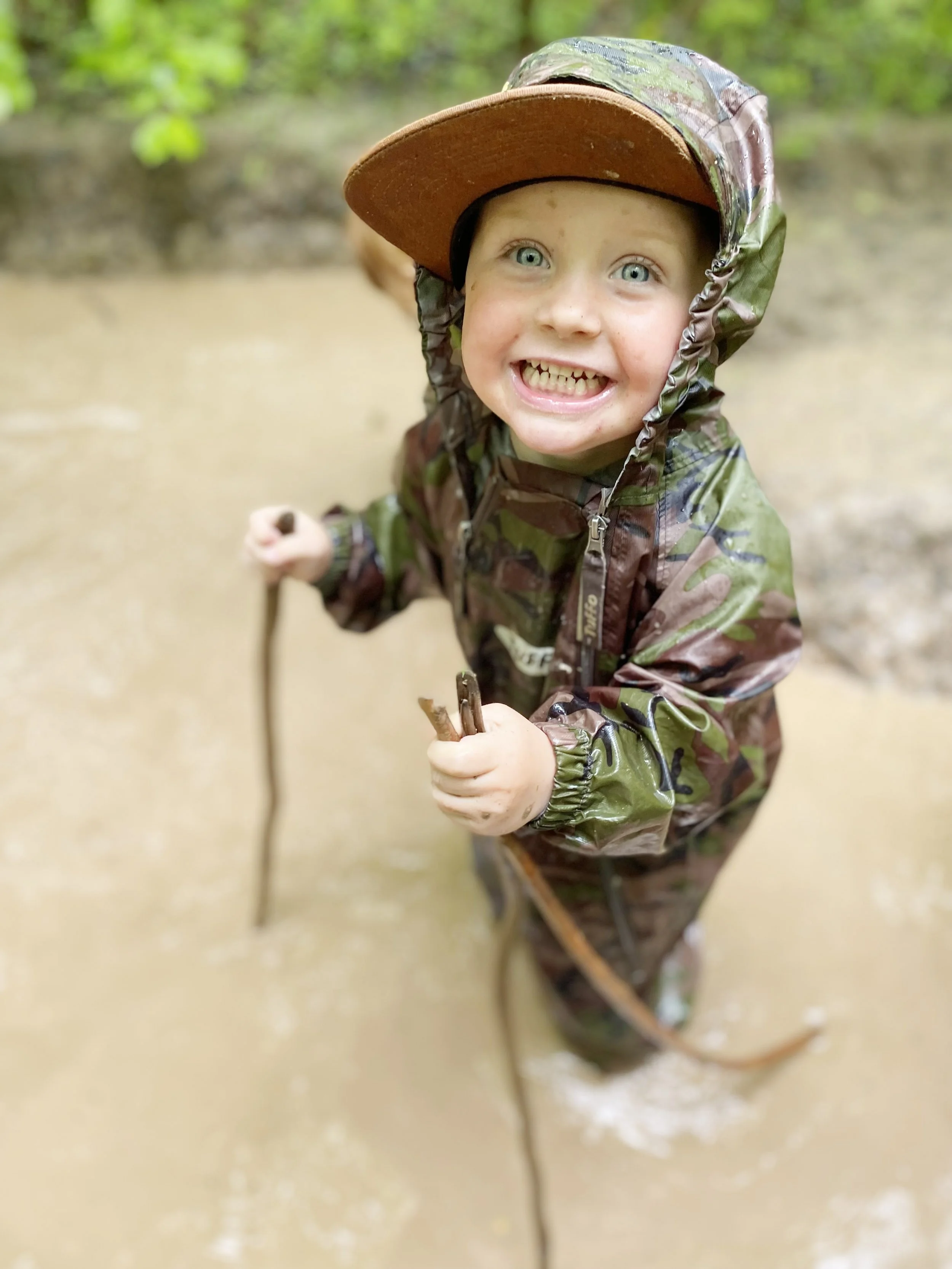 A young boy with blue eyes and a big smile, dressed in a camouflage waterproof jacket and hat, standing in a muddy outdoor area holding a stick.