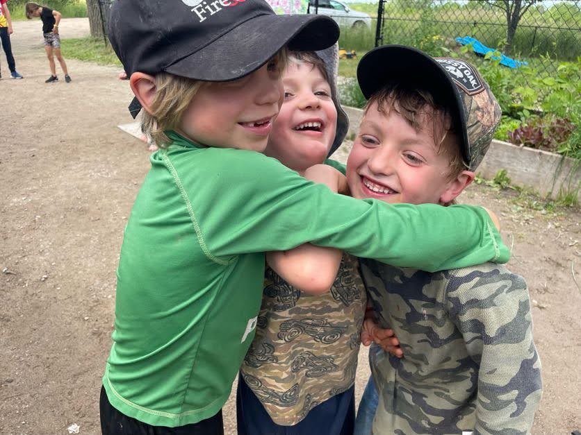 Three young boys are hugging each other and smiling outside on a dirt ground, with a fence and some plants in the background.