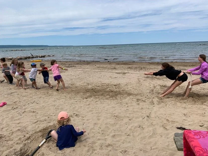 Children playing tug-of-war on a sandy beach near the ocean, with some kids sitting on the sand watching.