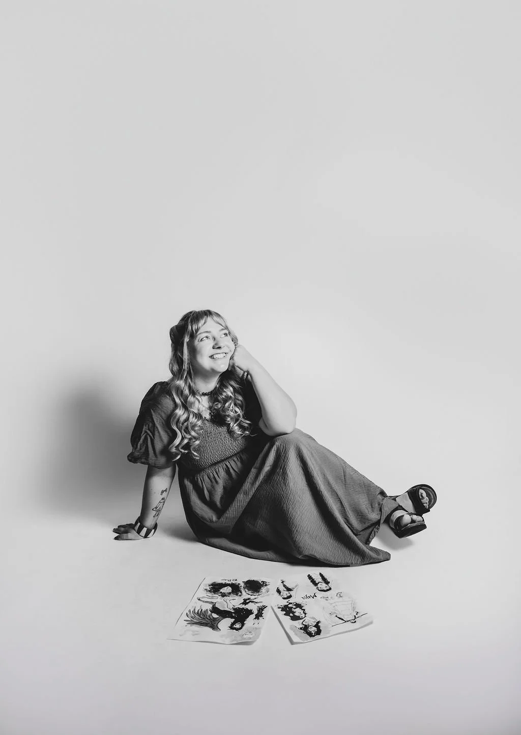 Black and white photo showing a woman with long wavy hair, smiling widely and looking up. She's wearing a long dress and is sitting on the ground. In front of her are laid out two sheets of paper with ink drawings.
