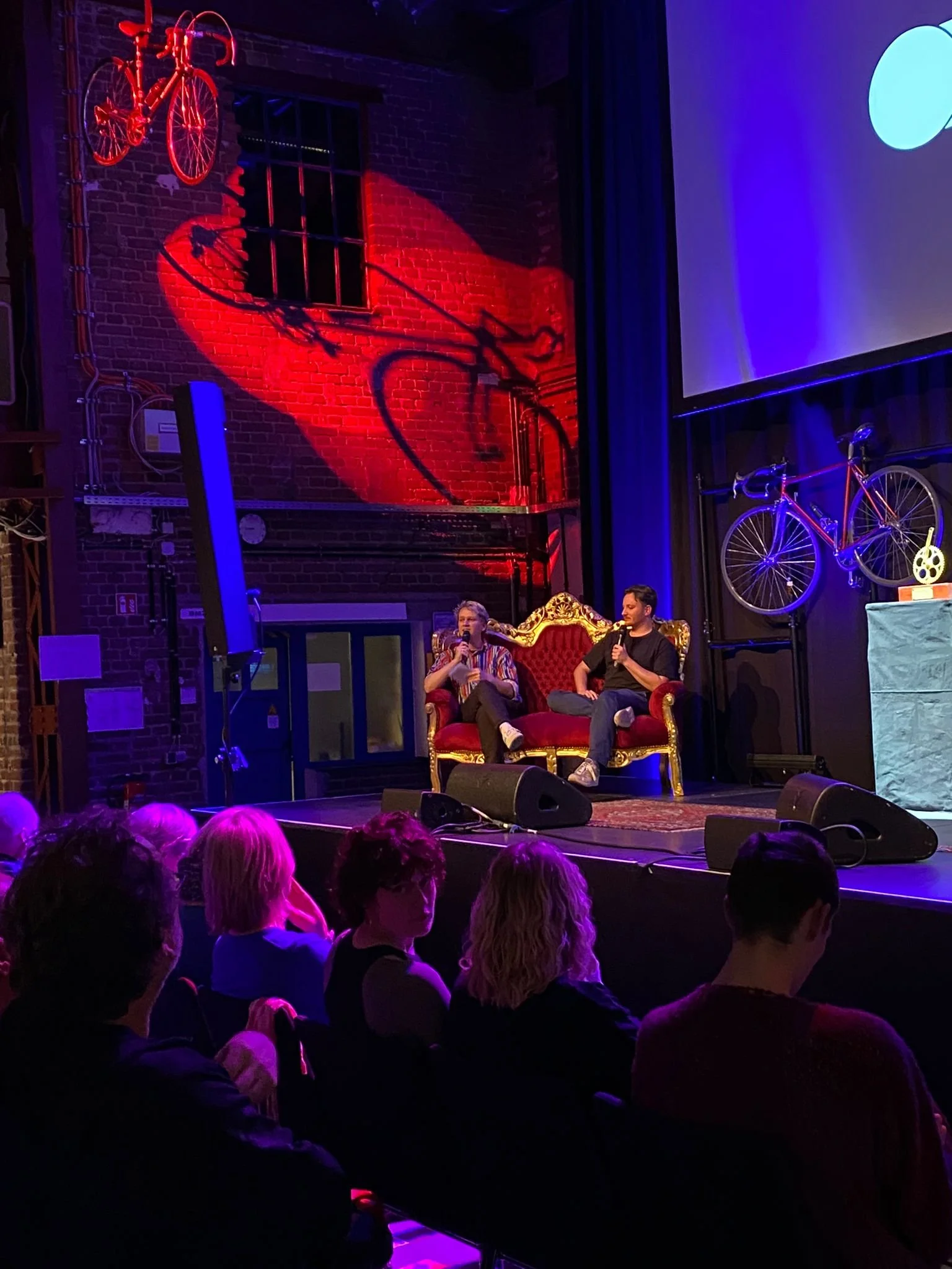 Two people sitting on a red and gold ornate couch on stage speaking into microphones, with a small audience seated in front under colorful stage lighting, behind them are two bicycles mounted on the wall and a large screen.
