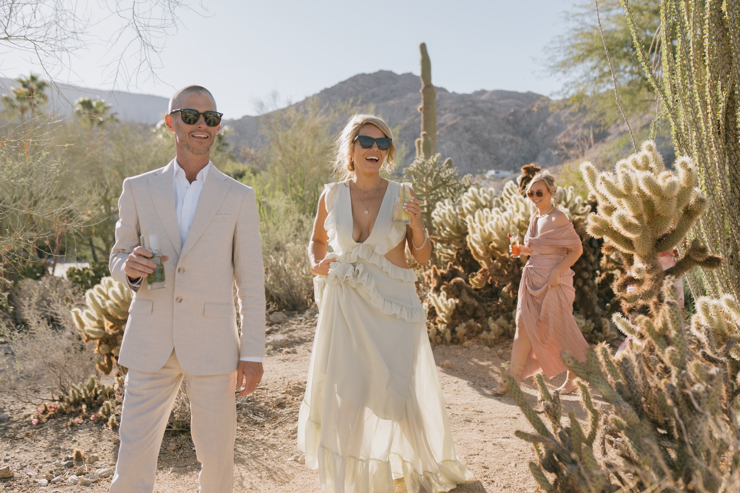 Group of people enjoying drinks at a desert outdoor gathering, with cacti and mountains in the background, dressed in light-colored clothing.
