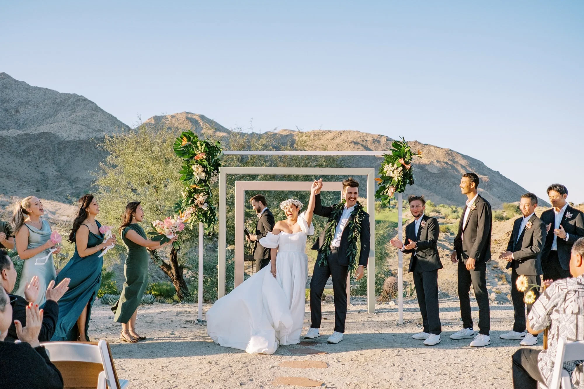 A wedding ceremony taking place outdoors in a desert landscape with mountains in the background. The bride and groom are celebrating, surrounded by bridesmaids and groomsmen, with guests clapping and cheering.