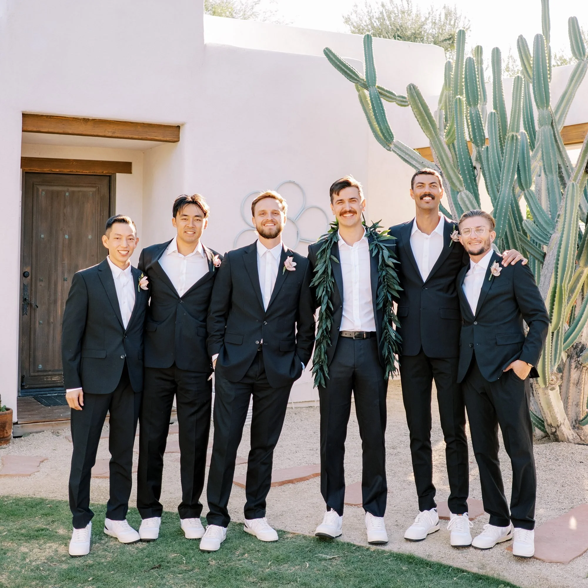A group of seven men in suits standing outdoors in front of a cactus and modern building, celebrating at a wedding.