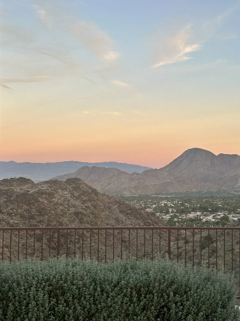 A scenic view of brown mountains and a valley at sunset, with a pastel-colored sky and a railing in the foreground.