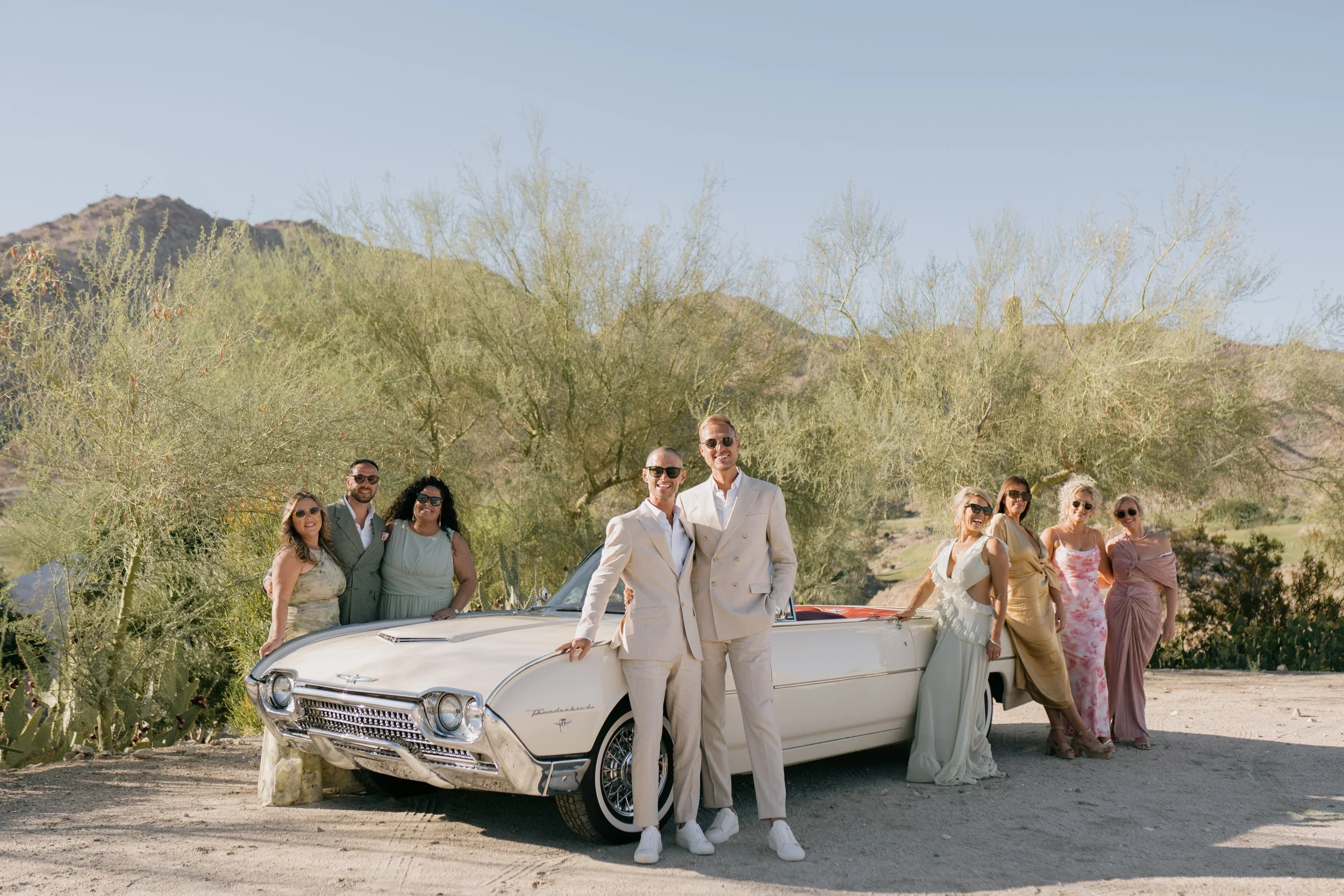 Group of people in formal attire posing next to a vintage white convertible car in a desert landscape with mountains and sparse trees in the background.