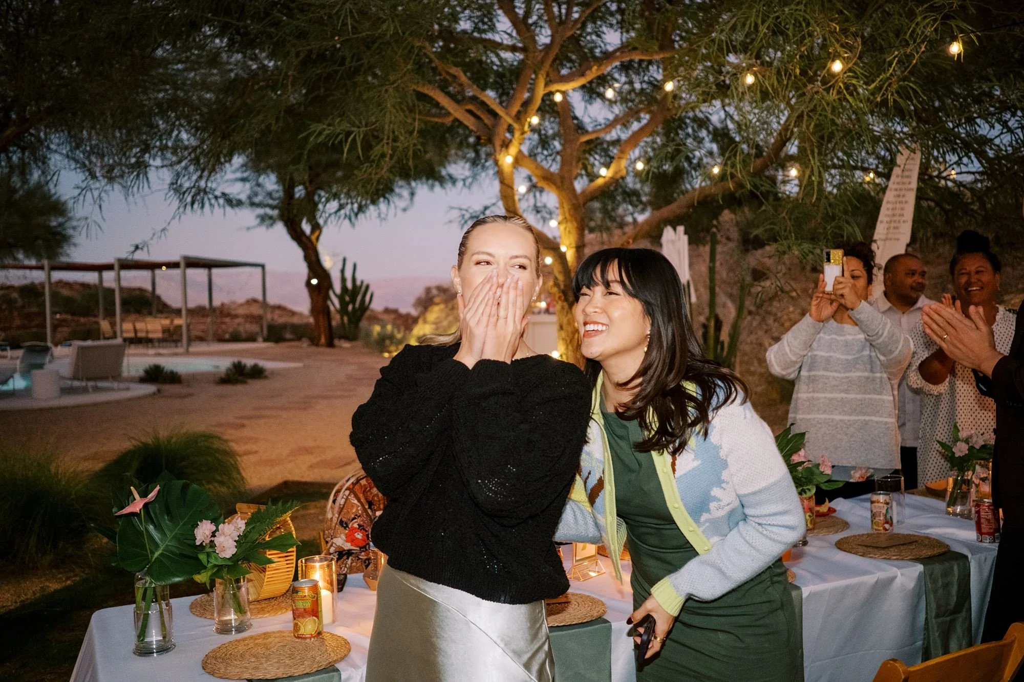 Two women are smiling and laughing together at an outdoor gathering during the evening, surrounded by friends, with a decorated table and string lights hanging from trees in the background.