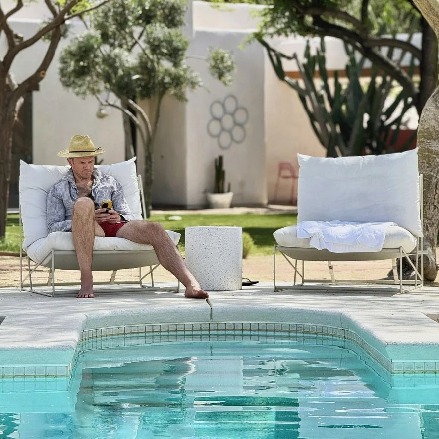 A man sitting on a poolside lounge chair using a smartphone, wearing a straw hat, a striped shirt, and red shorts, with another empty lounge chair nearby, in a backyard with a swimming pool and trees.