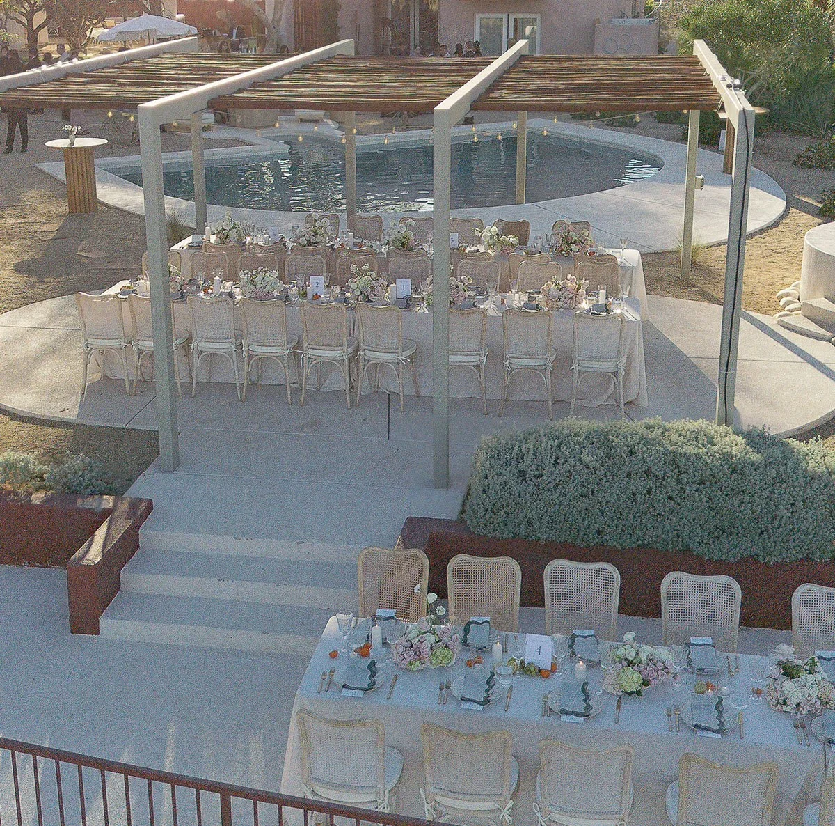 Outdoor wedding reception setup with long dining tables decorated with floral centerpieces, set beside a swimming pool with a pergola overhead.
