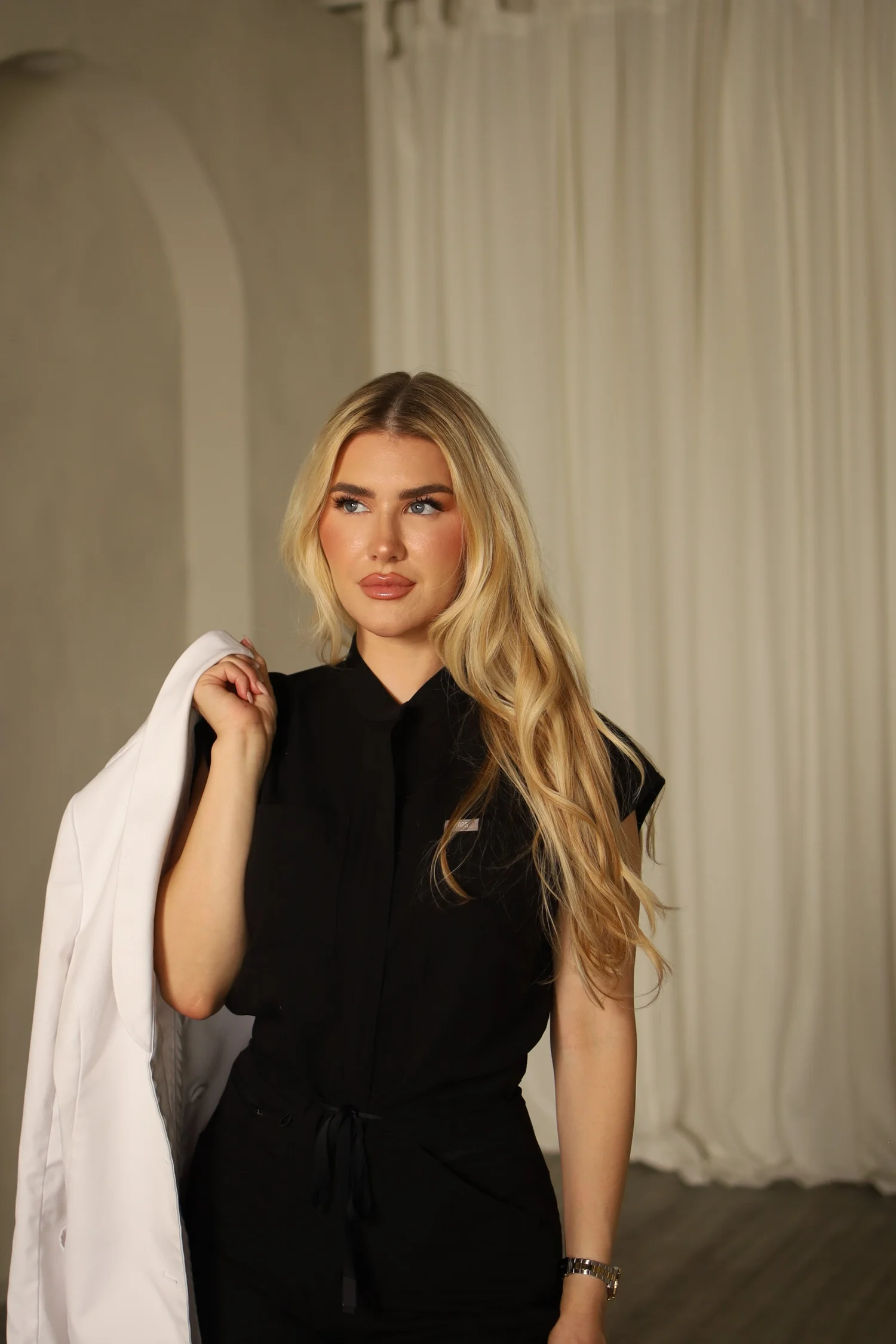 Dr. Alex, DNP, APRN, FNP-C, founder of Blonde Girl Aesthetics, in black scrubs with her white coat over her shoulder