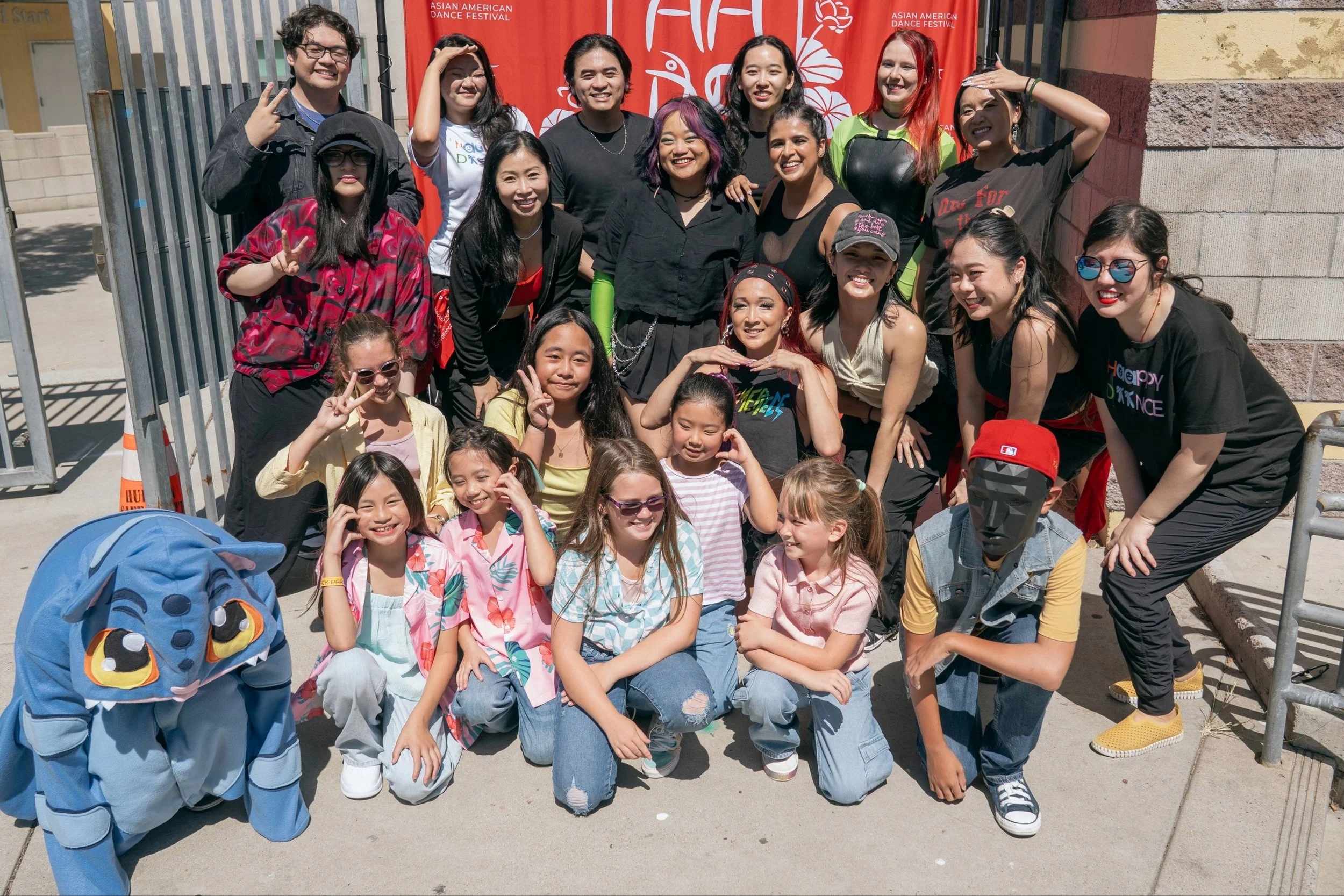 Group of people, including children and adults, smiling and posing for a photo outdoors in front of a red banner that reads 'Asian American Dance Festival'.