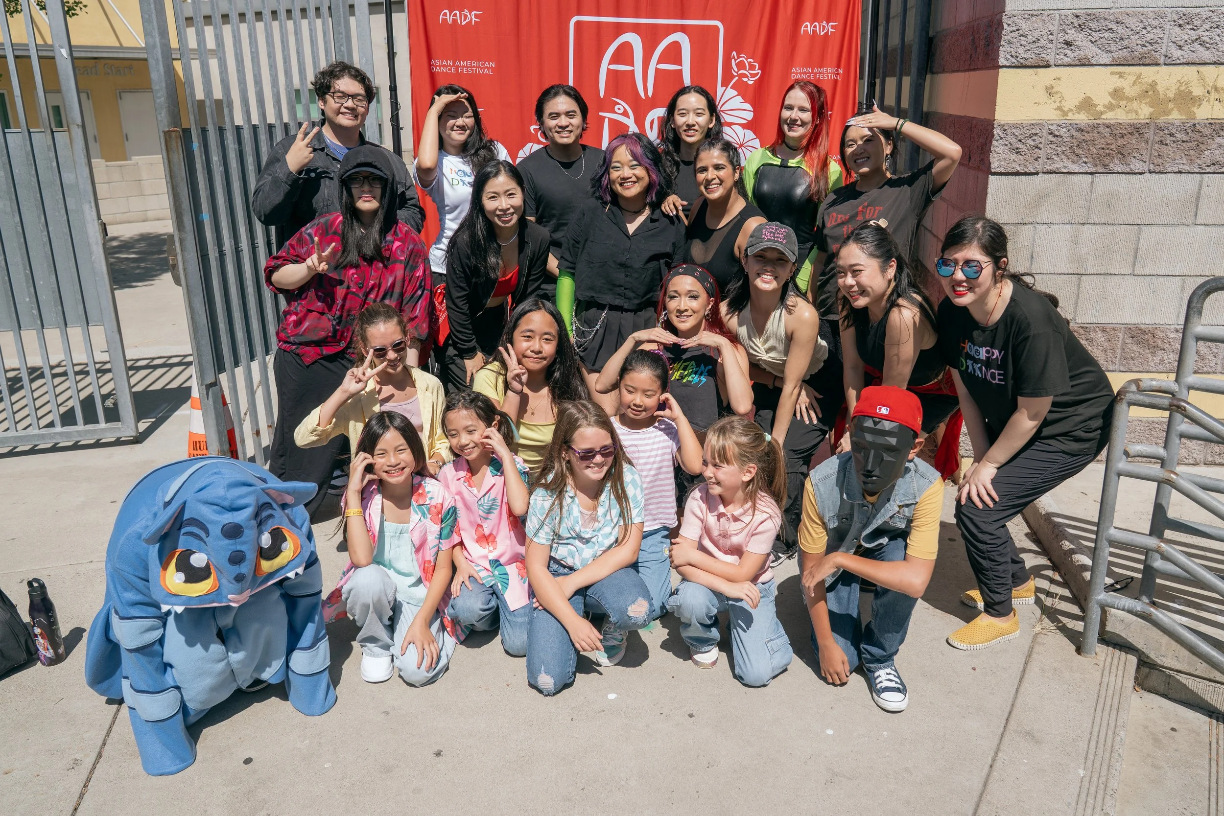 Group of children and adults posing outdoors in front of a red banner with white text and graphics at the Asian American Dance Festival.