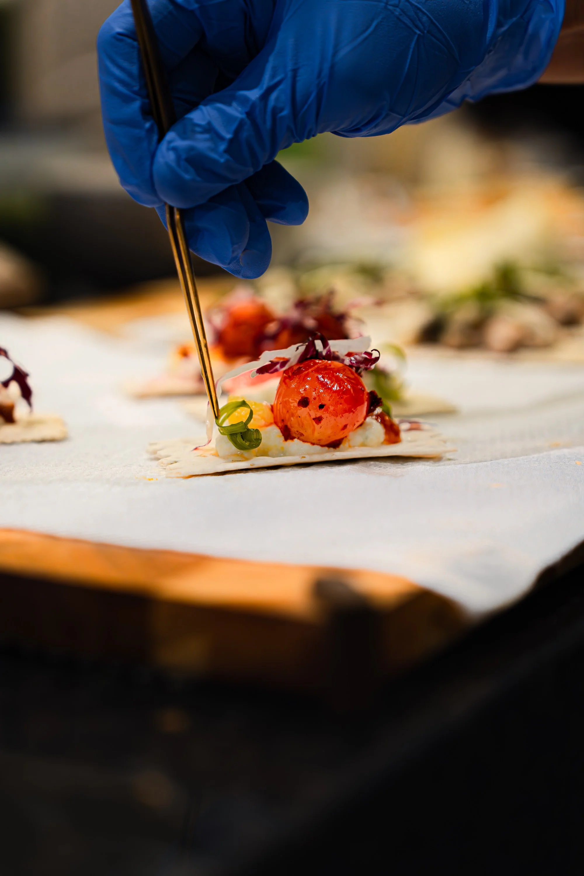 A chef wearing a blue glove uses tweezers to place a garnish on a small, detailed appetizer with colorful ingredients on a rectangular white plate.