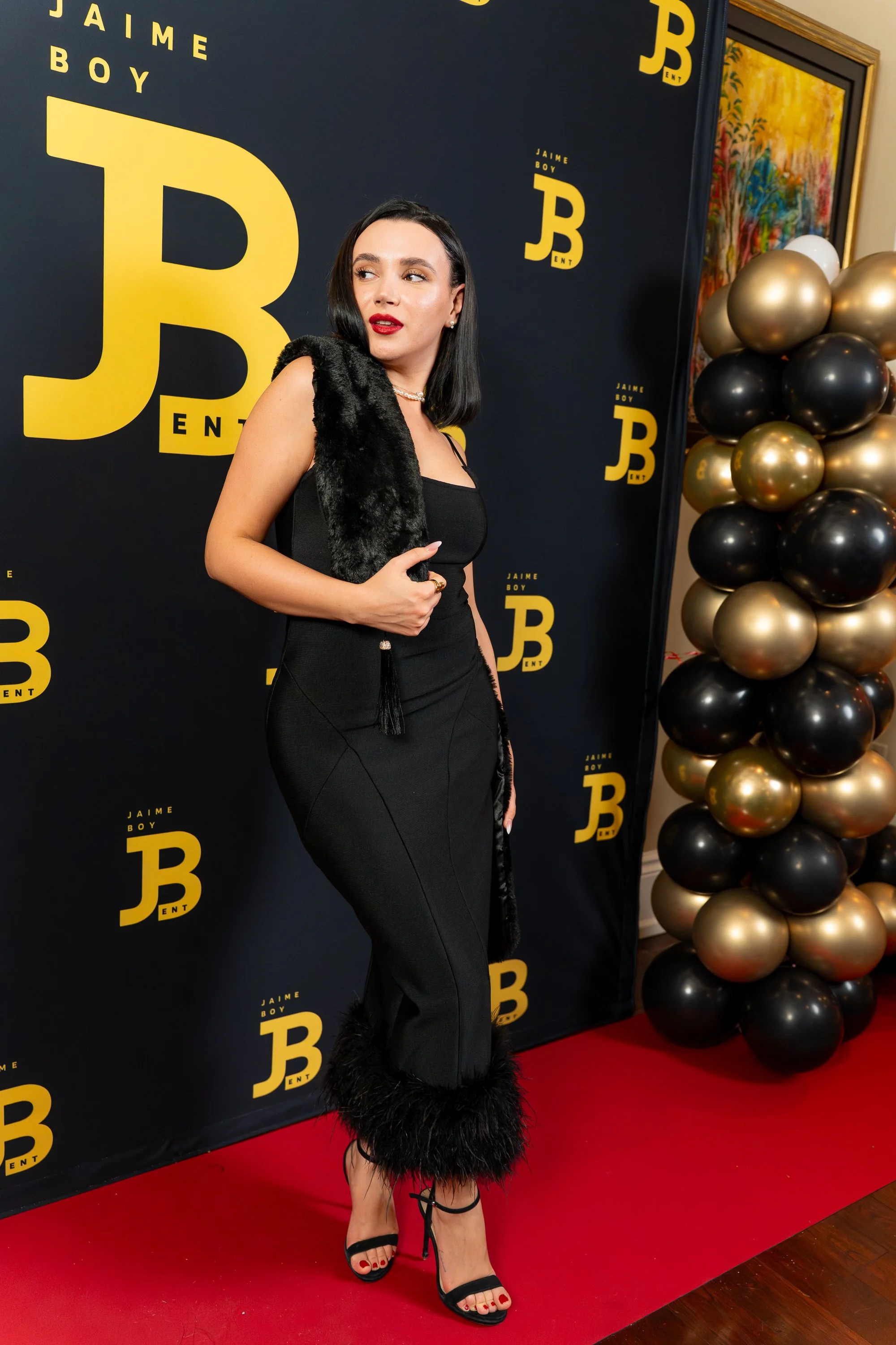 A woman dressed in a black dress with feathered accents around the hem and over her shoulders, wearing black heels with ankle straps, standing on a red carpet at a promotional event for Jaime Boy, with black and gold balloons and a black backdrop displaying the artist's name and logo.