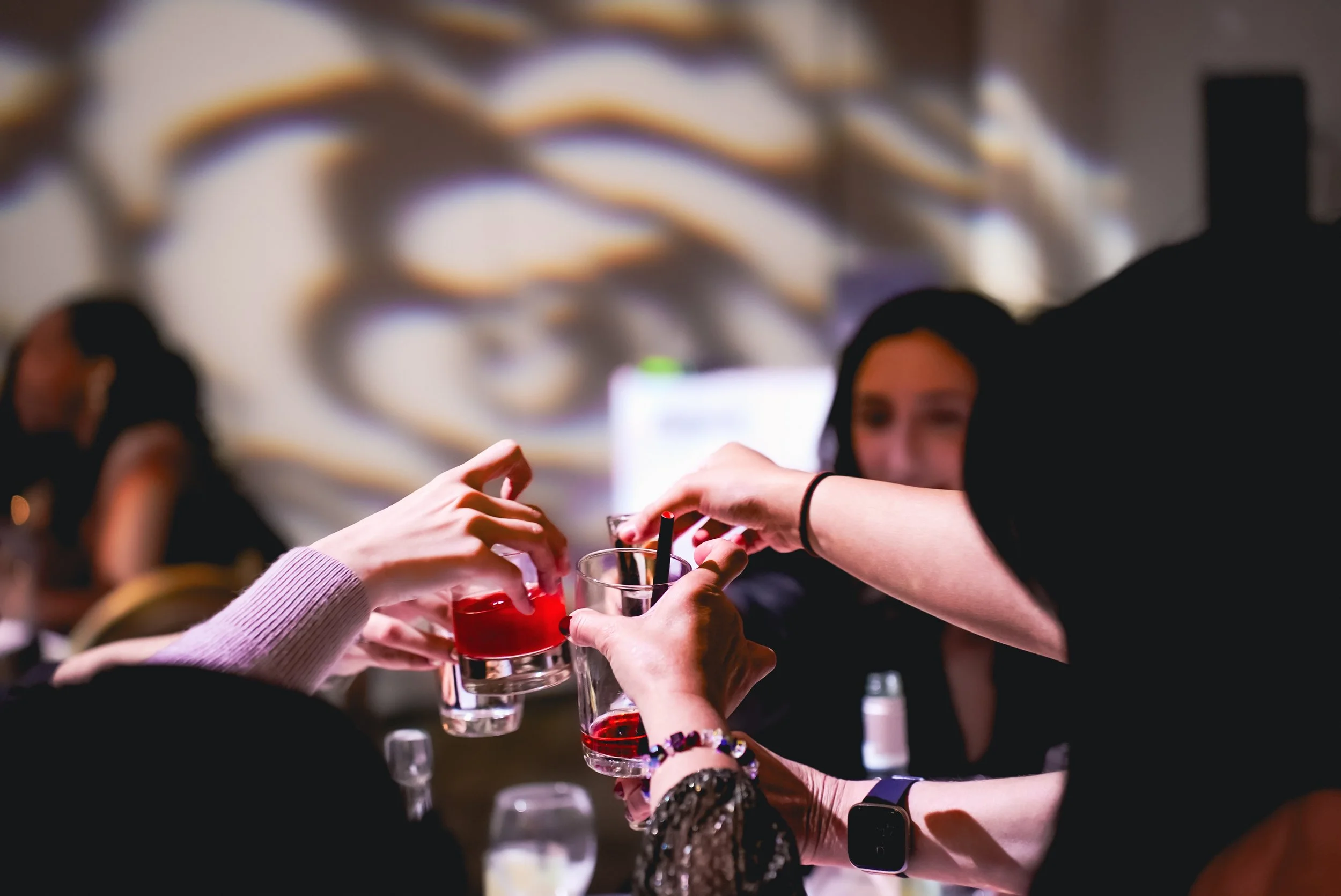 Group of friends sharing drinks at a social gathering, raising glasses of red beverage for a toast in a cozy indoor setting with abstract wall art in the background.
