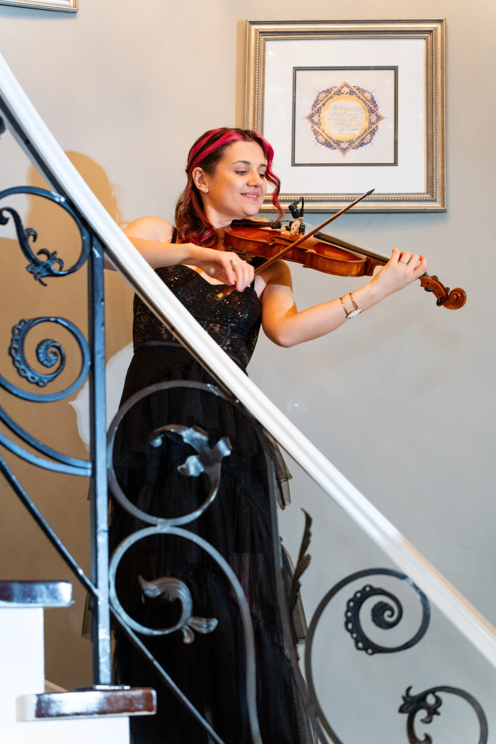 A woman with pink streaks in her hair playing a violin on an indoor staircase. She is wearing a black dress with a sparkly top and a long black skirt. There is a framed art piece with Arabic calligraphy on the wall behind her.