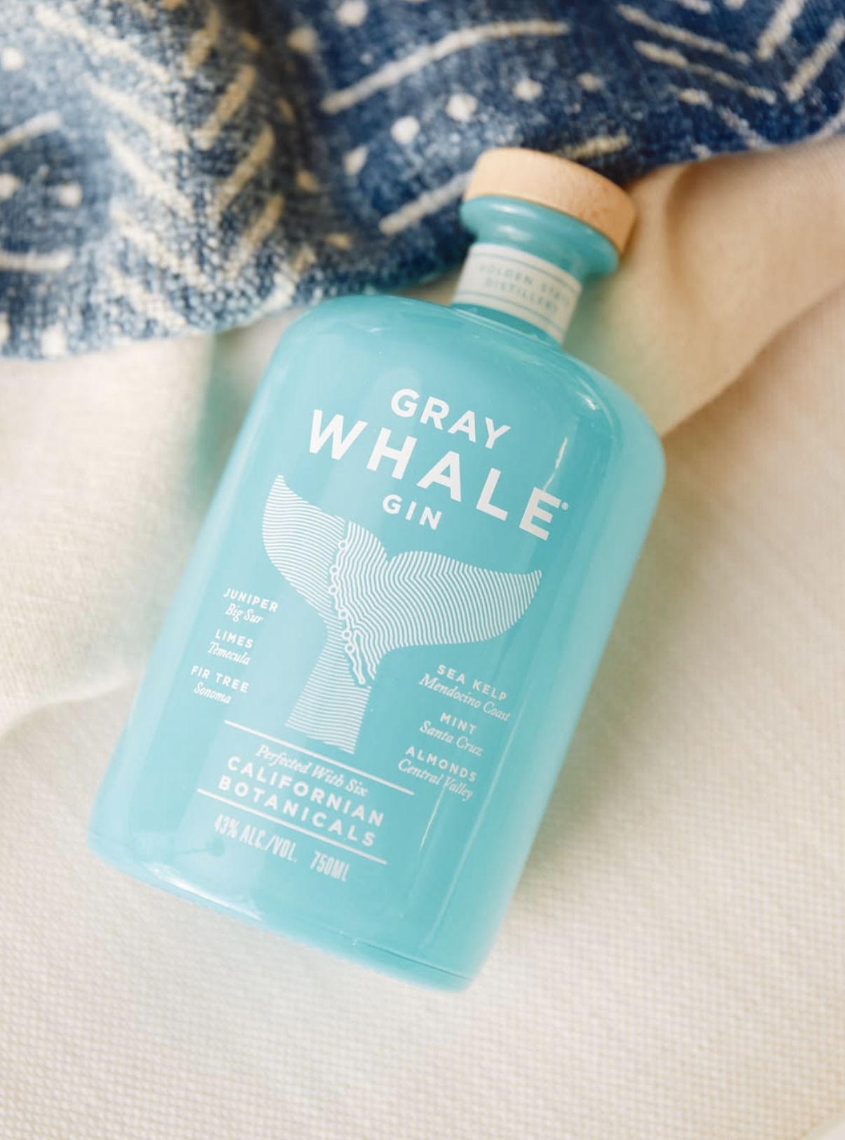Bottle of Gray Whale Gin resting on sand beside a blue patterned towel, highlighting its turquoise glass and whale tail label design.
