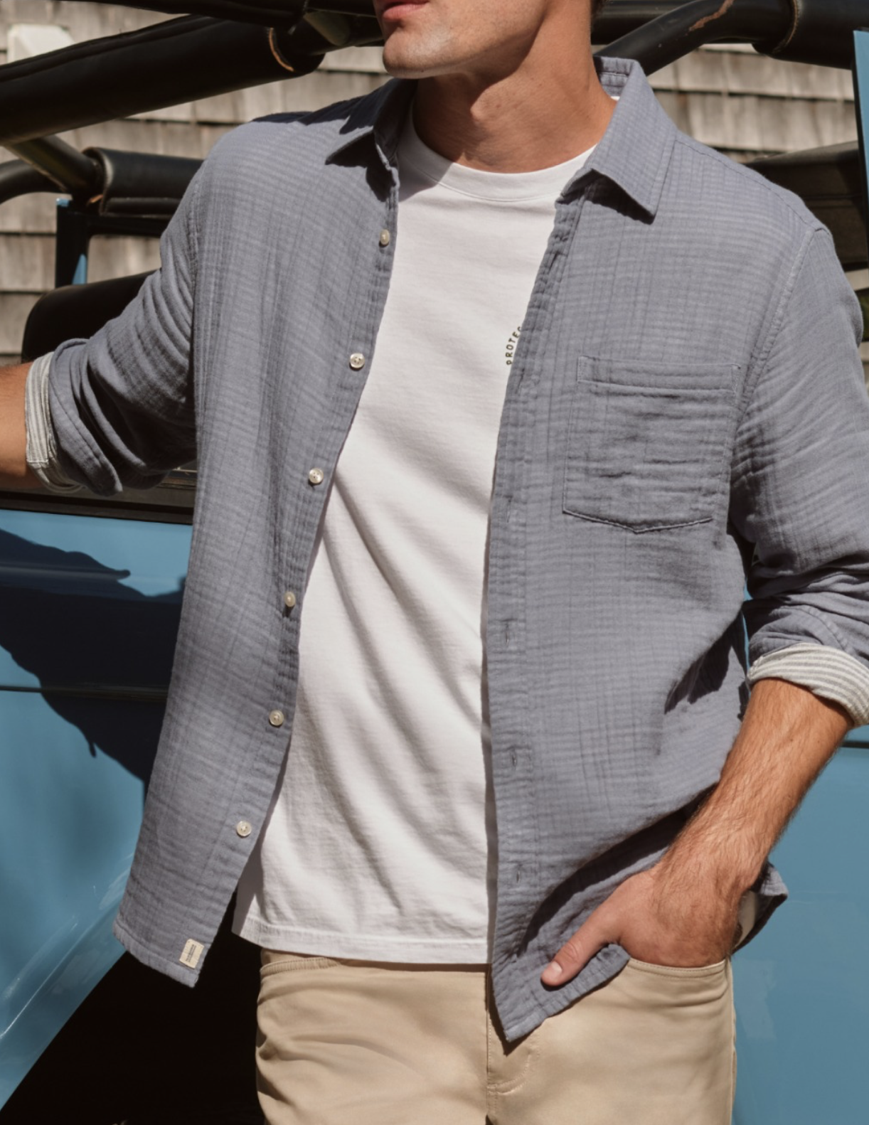 Man wearing a light gray textured button-down shirt over a white tee with khaki pants, leaning against a vehicle in a casual outdoor setting.