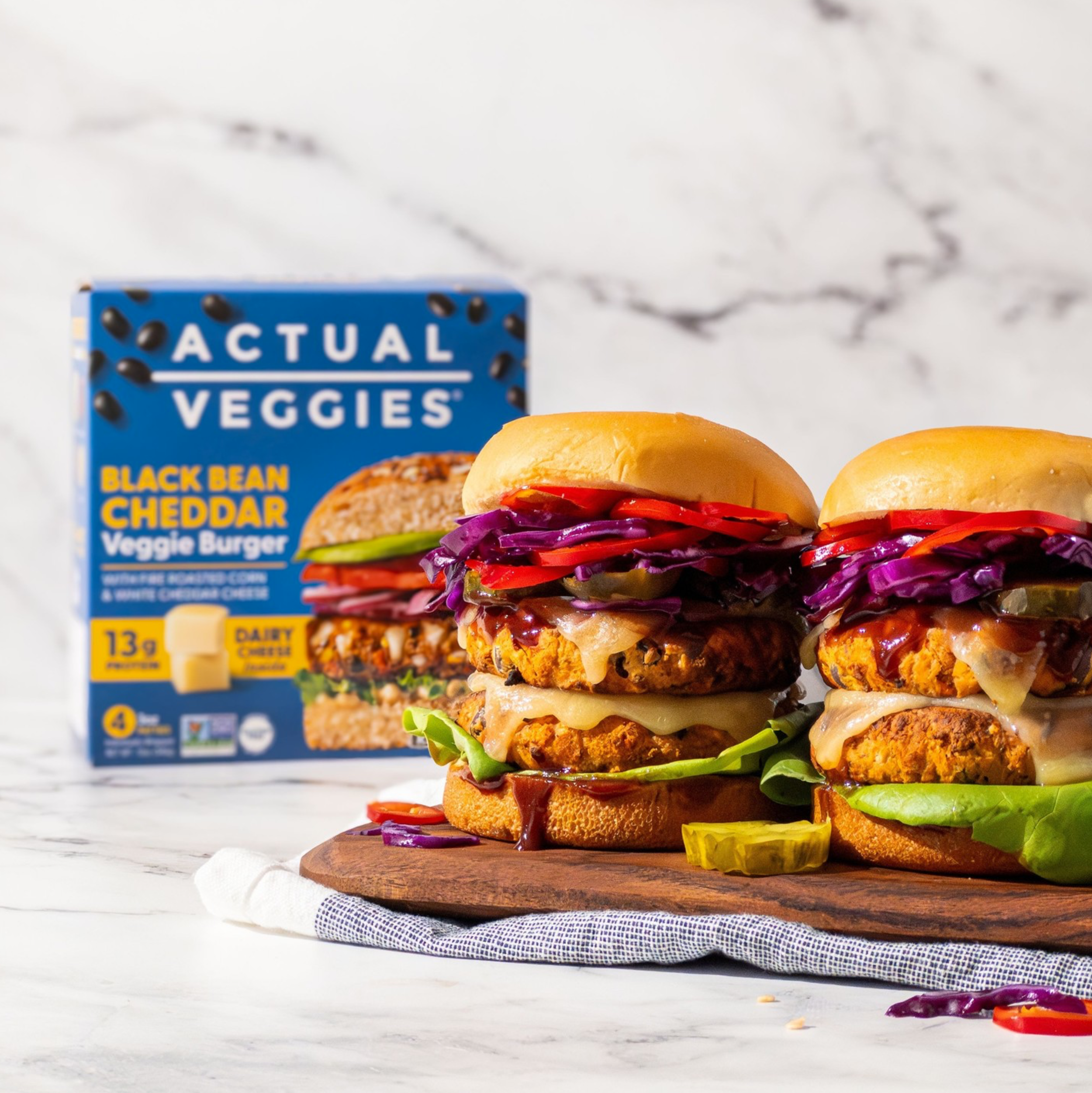 Two Actual Veggies Black Bean Cheddar Veggie Burgers with fire roasted corn and white cheddar cheese, topped with red cabbage, roasted peppers and pickles on a wooden cutting board.