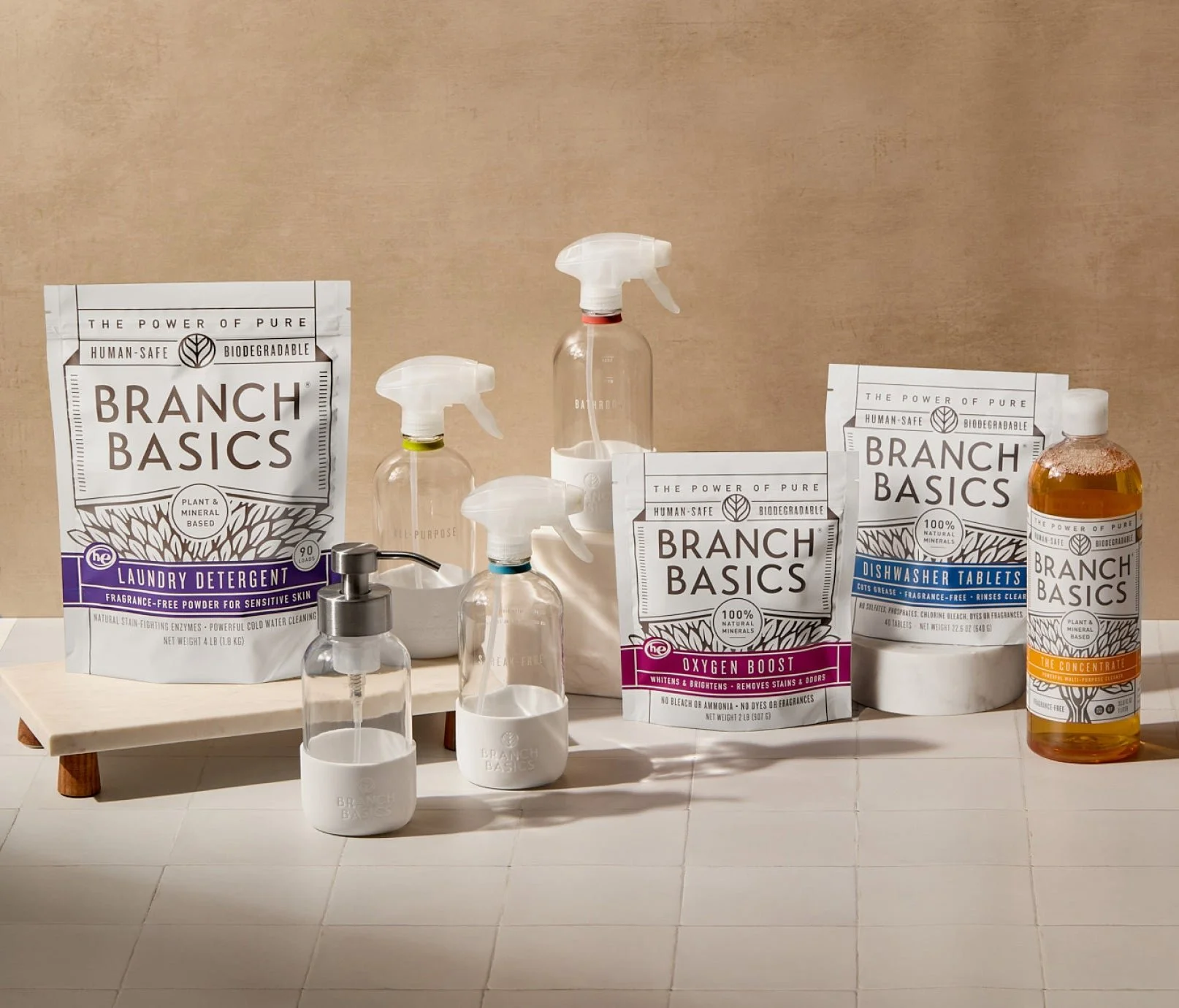 Branch Basics cleaning products arranged on a neutral countertop, including refill pouches, a concentrate bottle, spray bottles and foaming hand soap dispenser, showcasing a simplified, human-safe home cleaning system