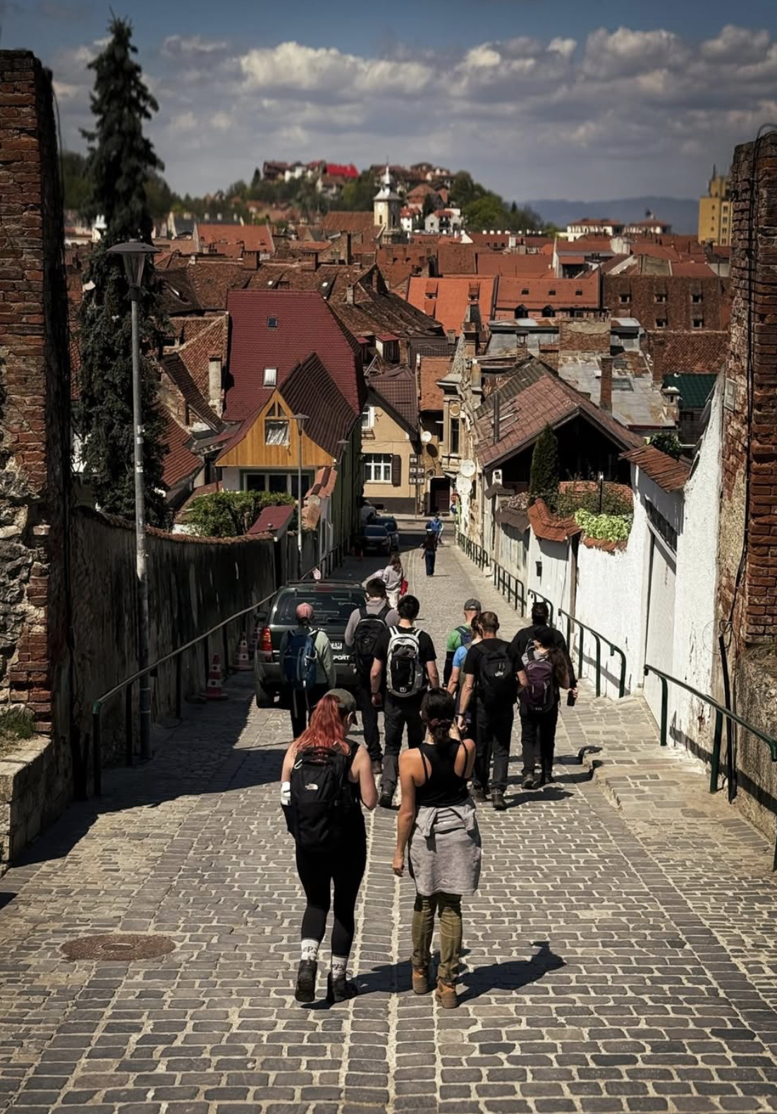 Group of travelers walking down a cobblestone street in a historic European town, with colorful rooftops and buildings stretching across the hillside in the background