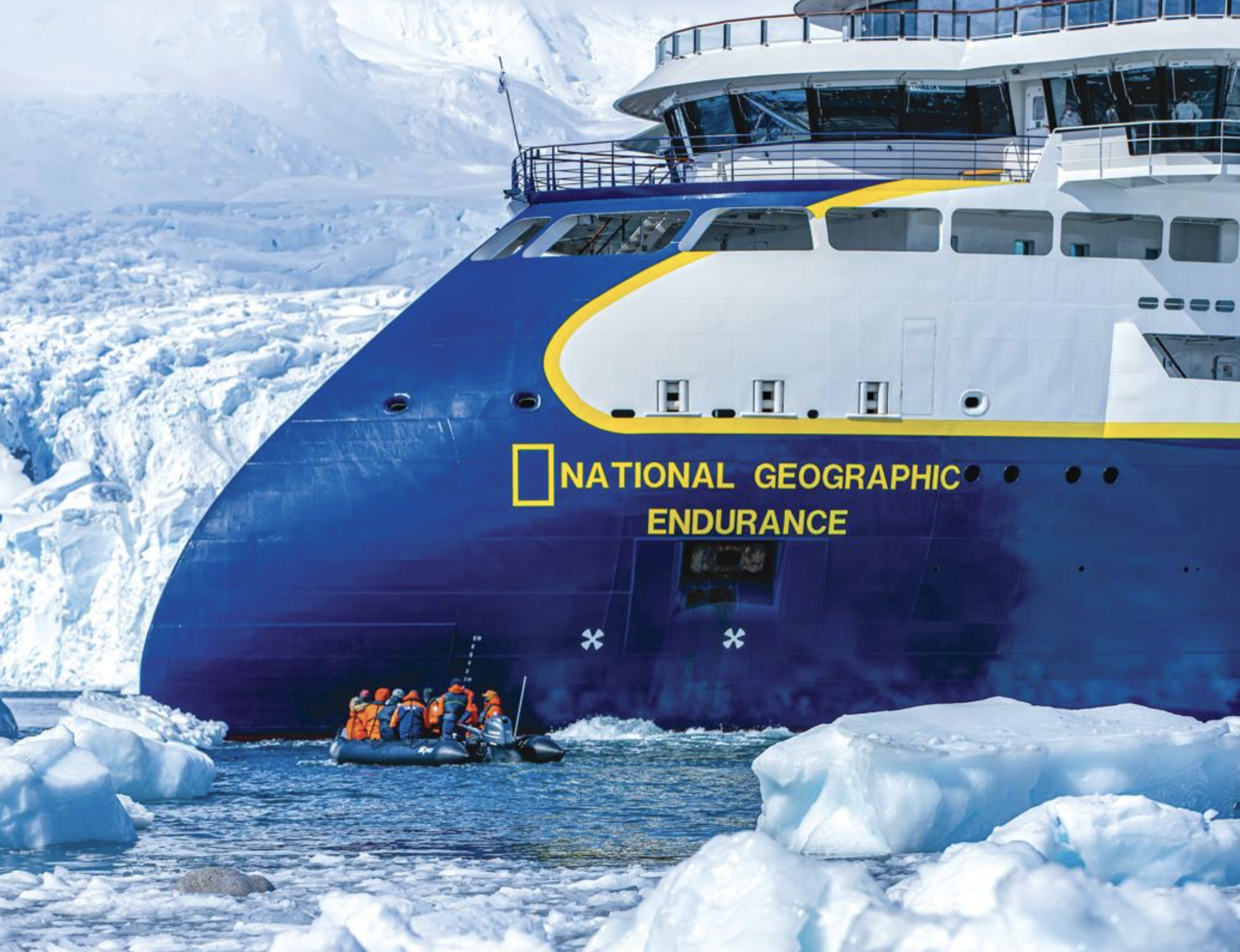 National Geographic Endurance expedition ship navigating icy Antarctic waters as a small zodiac boat with passengers approaches floating sea ice and glaciers nearby.