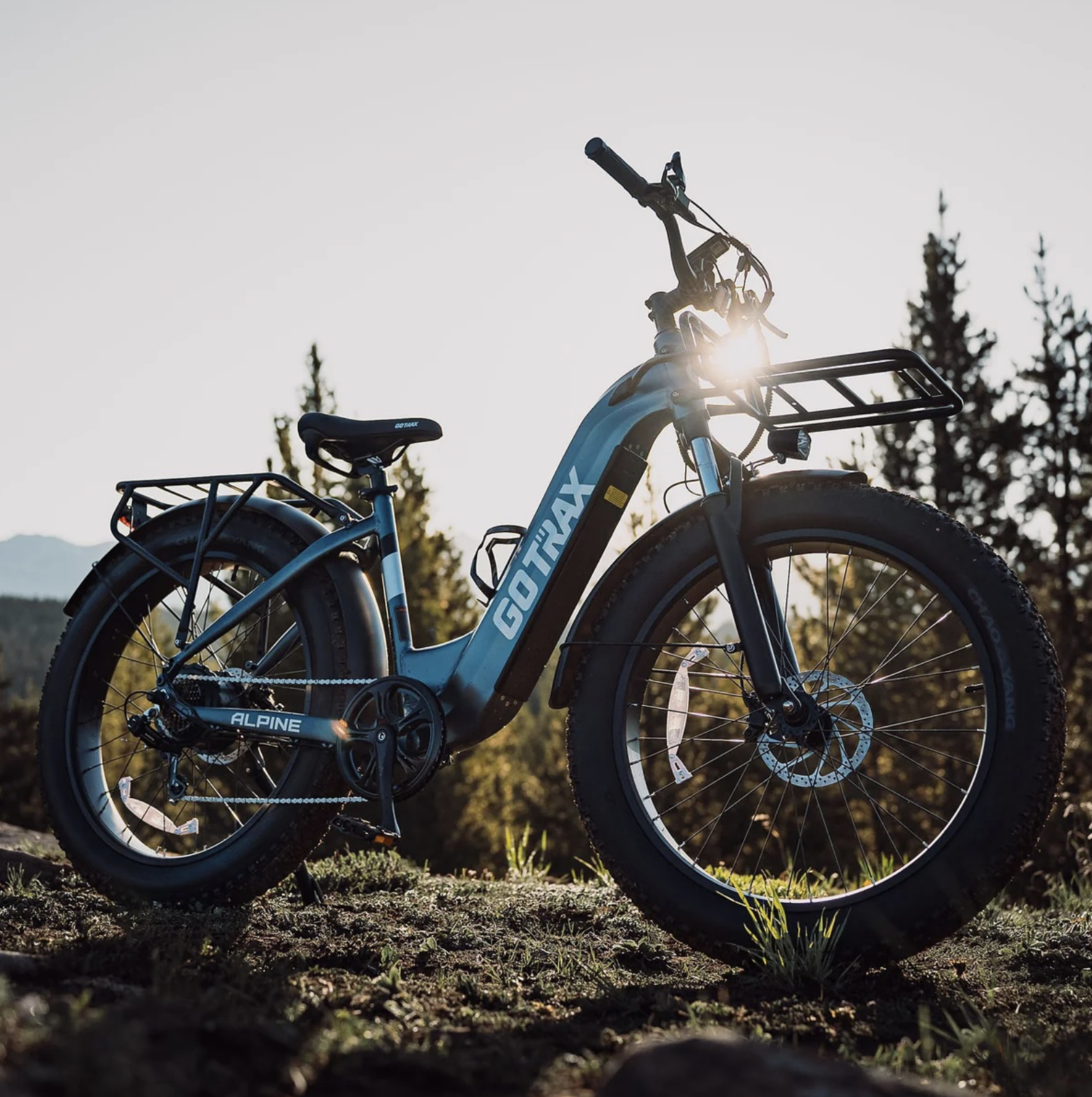 Electric fat-tire GOTRAX Alpine e-bike parked on a dirt trail with trees and mountains in the background during golden hour.