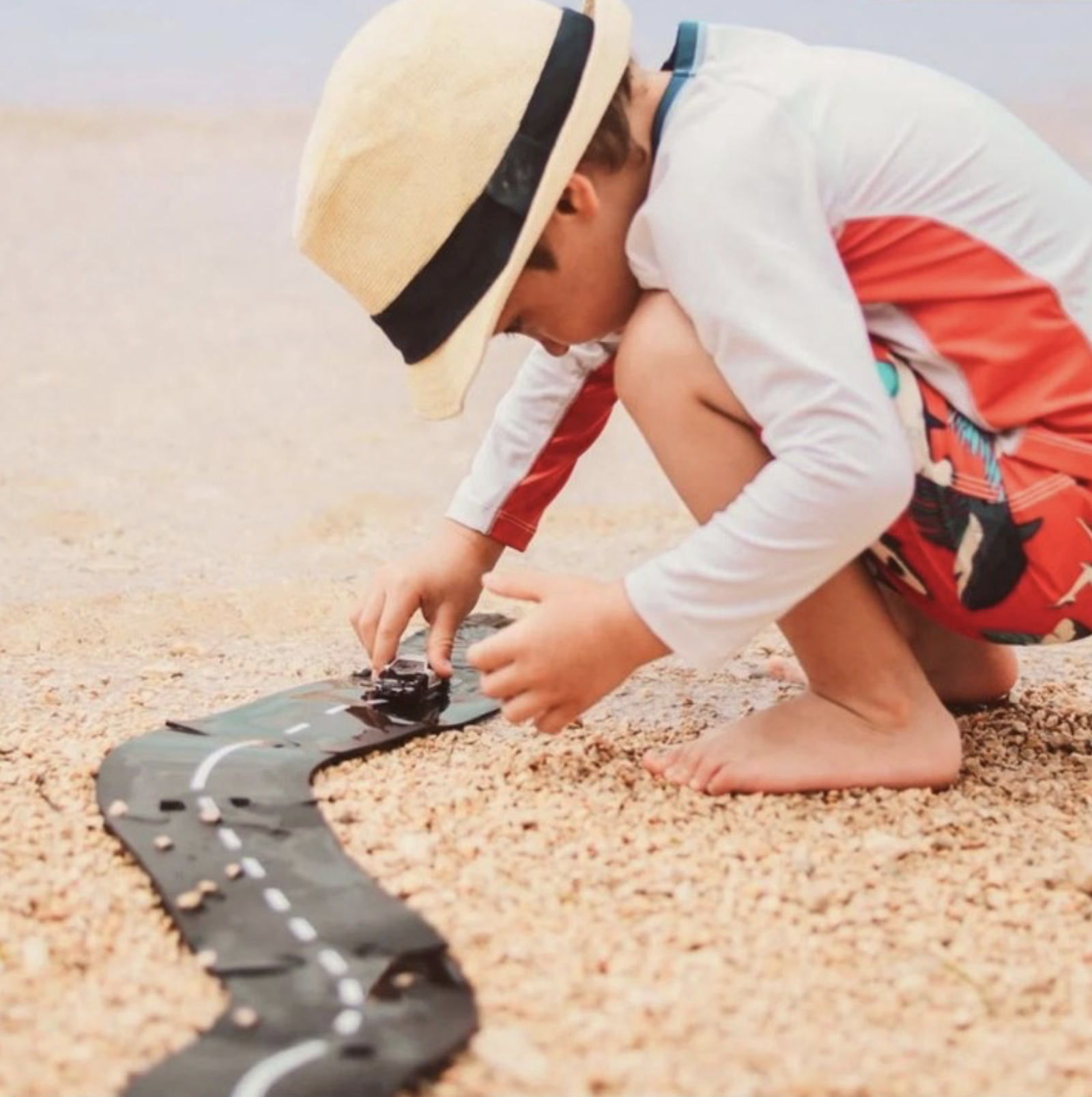 Child crouching on a sandy beach, playing with a toy road track and small car, wearing a sun hat and long-sleeve top while focused on imaginative play outdoors.