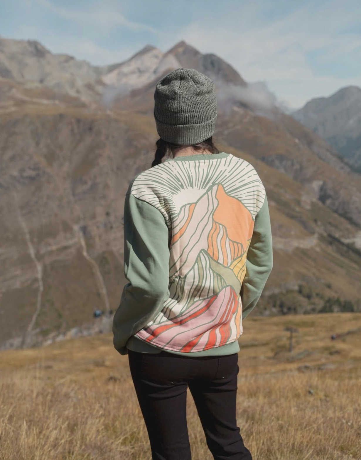 Person wearing a green sweatshirt with an abstract mountain graphic on the back and a knit beanie, standing on a grassy hillside overlooking a rugged mountain landscape.